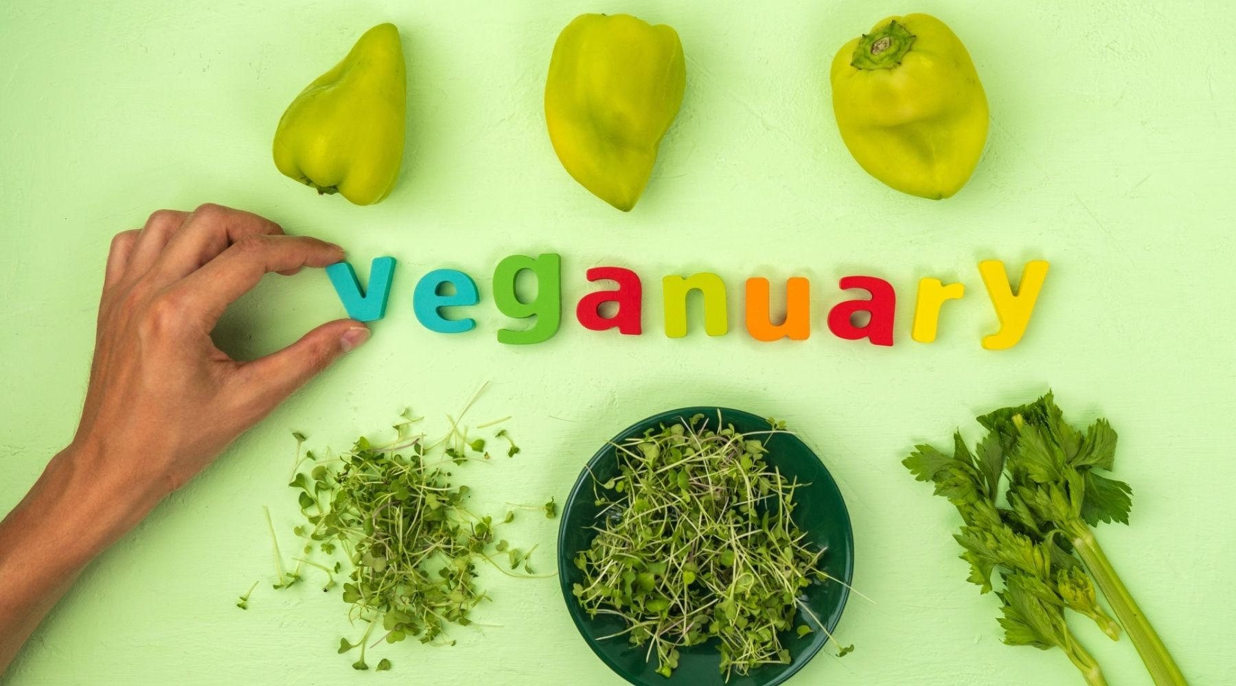 A photo flatly on a light green background. 'Veganuary' is spelled out in colourful fridge magnet letters, with a hand adjusting the 'v'. There are green peppers above the letters and green herbs below.