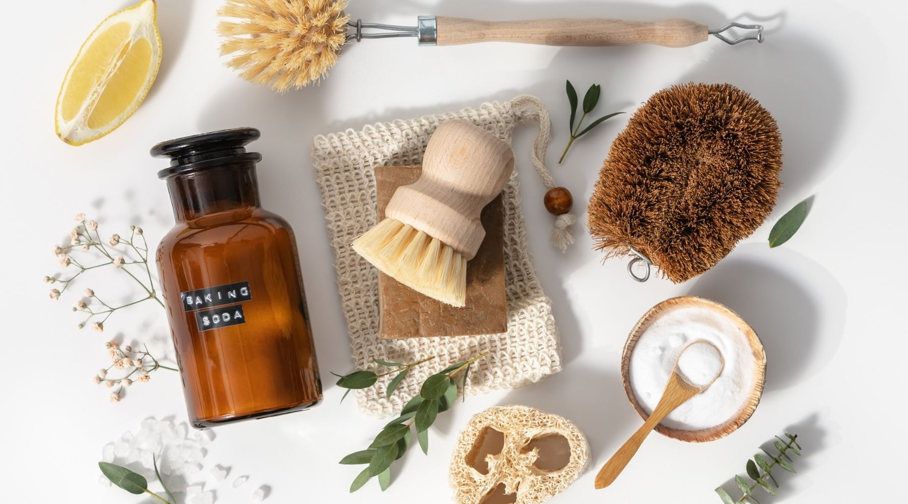 Flat lay photo of an array of natural cleaning items: a coconut scrubbing pad, wooden brush, net soap bag, loofah, lemon slice, and bowl of baking soda.