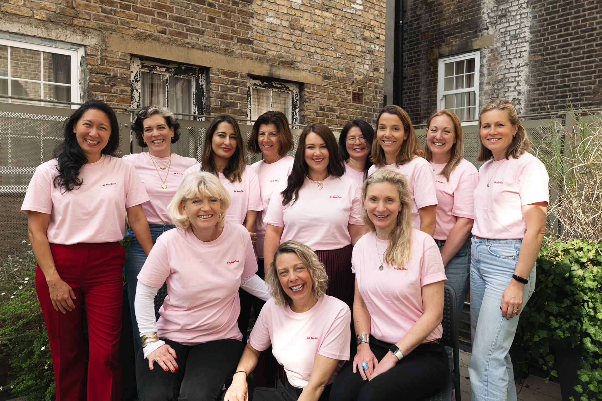 Photo image showing 12 women smiling and posing together outside in a garden with a building in the background. All the women are wearing pink t shirts with 'Be Fearless' embroidered in red thread on the left hand side.