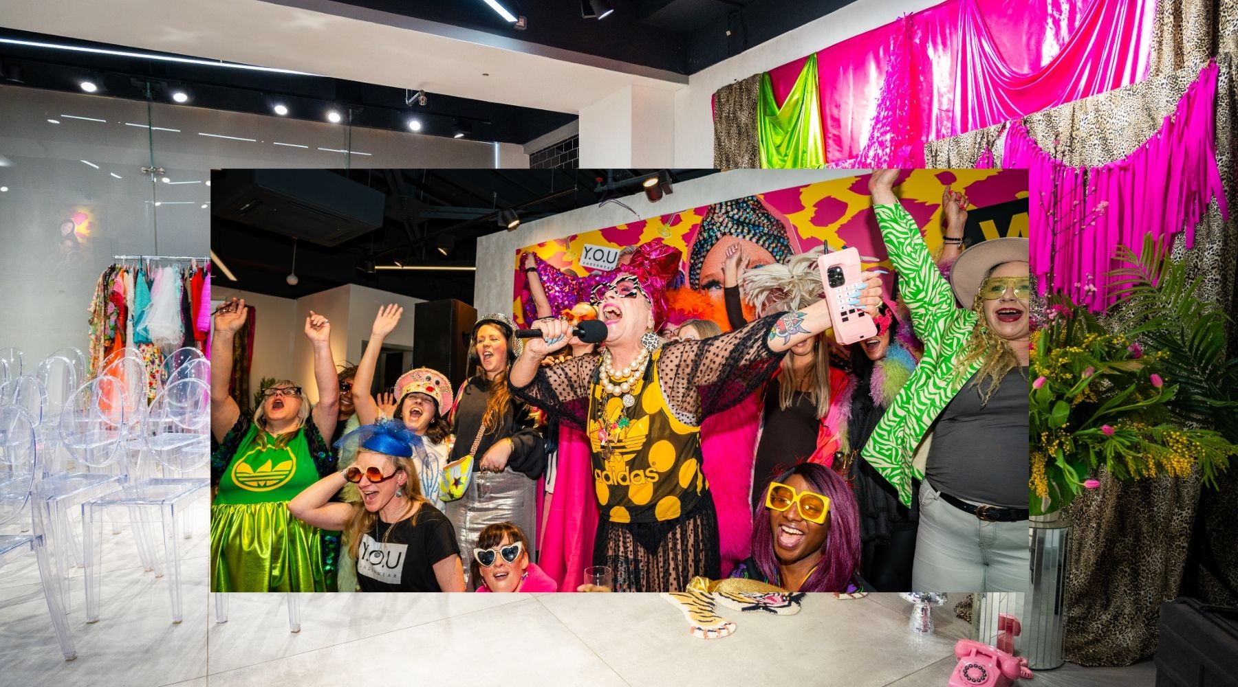 Colourful photo of a group of brightly dressed women posing together with their arms in the air against a photo of the inside of a venue.