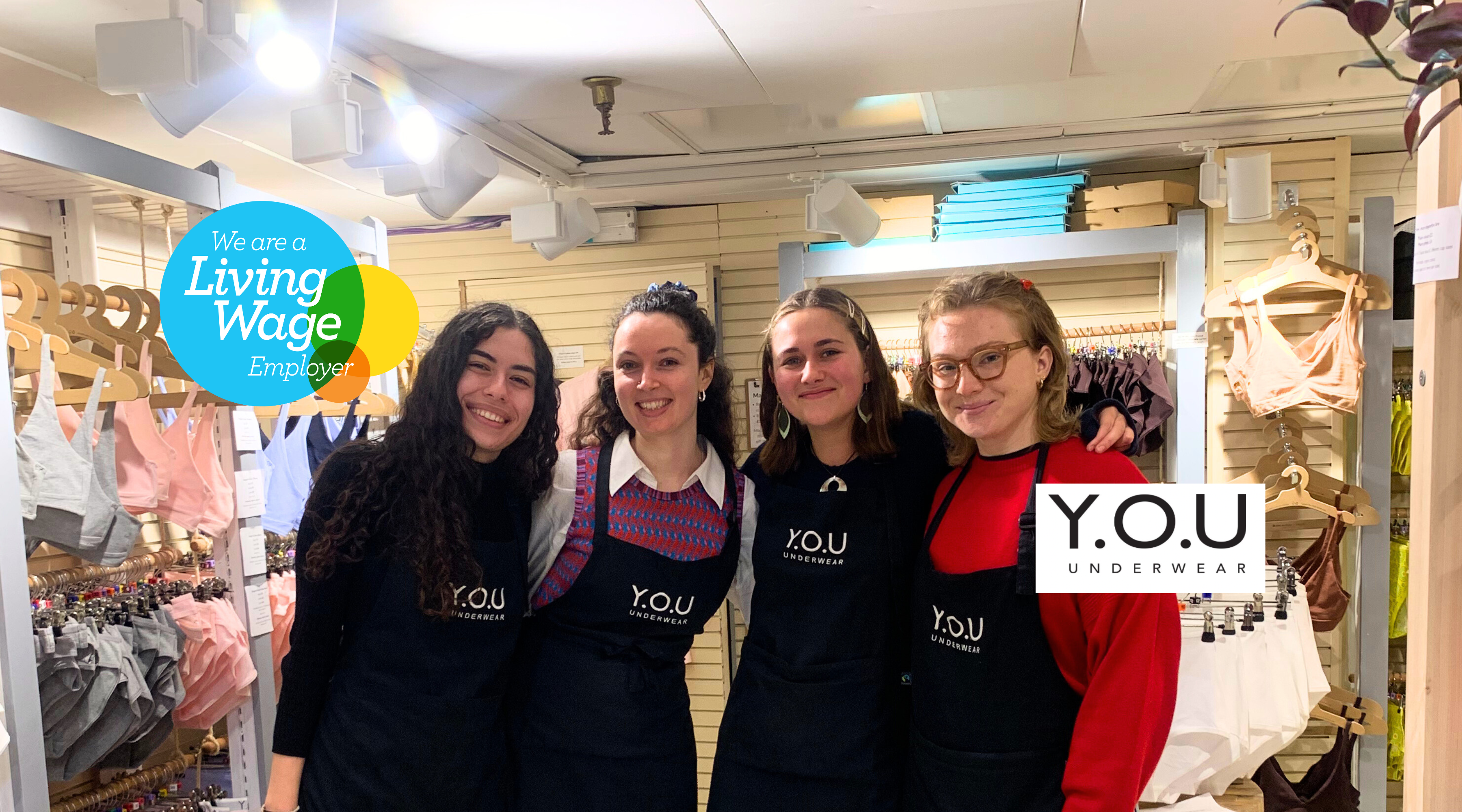 Photo showing four young women of similar height standing and posing together in front of an underwear display in a shop. Over the image is the Living Wage logo and the Y.O.U logo