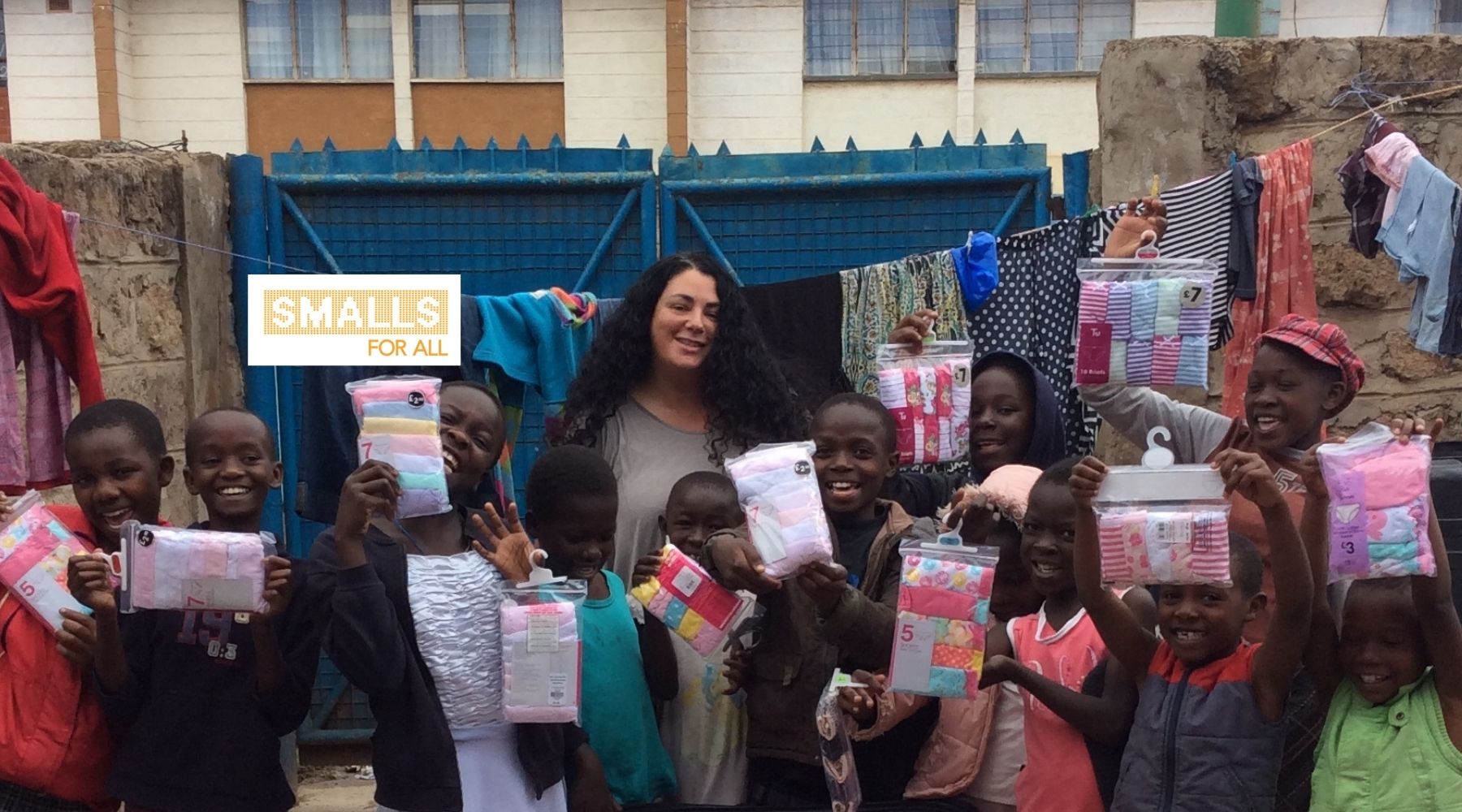 Photo of a woman with long dark hair standing with a group of children who are all smiling towards the camera and holding up package of underwear from donations. The white and yellow Smalls For All logo is laid over the top of the photo./