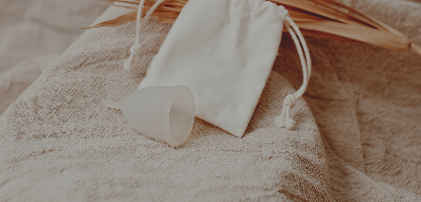 A clear silicone menstrual cup is in the bottom middle of the screen, on its side. An organic cotton drawstring bag is behind it and behind that is a brown coloured leaf 