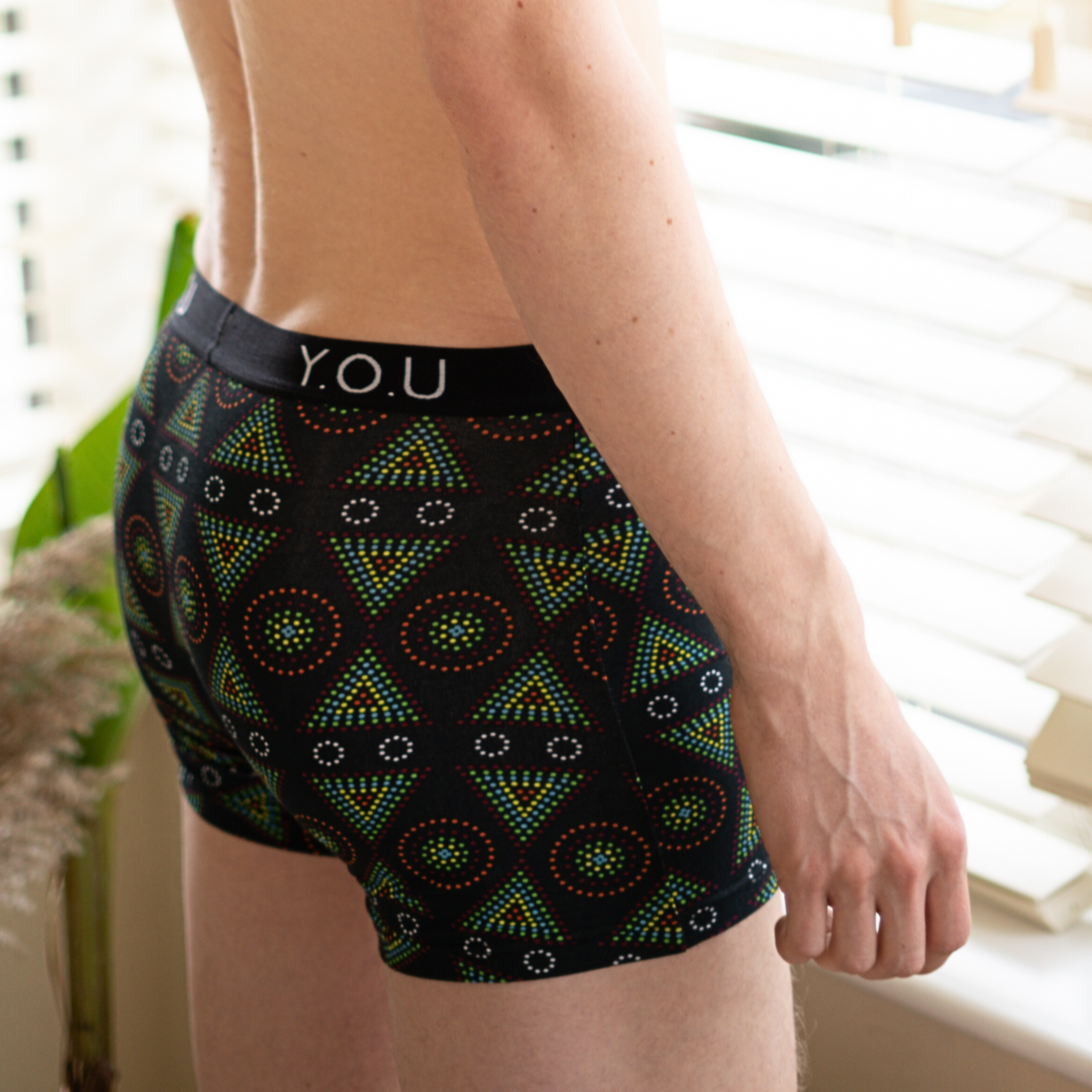A person wearing black hipster trunks with a colorful geometric pattern stands near a window with white blinds. The waistband of the underwear displays the initials "Y.O.U." in white. Plants are partially visible in the background.