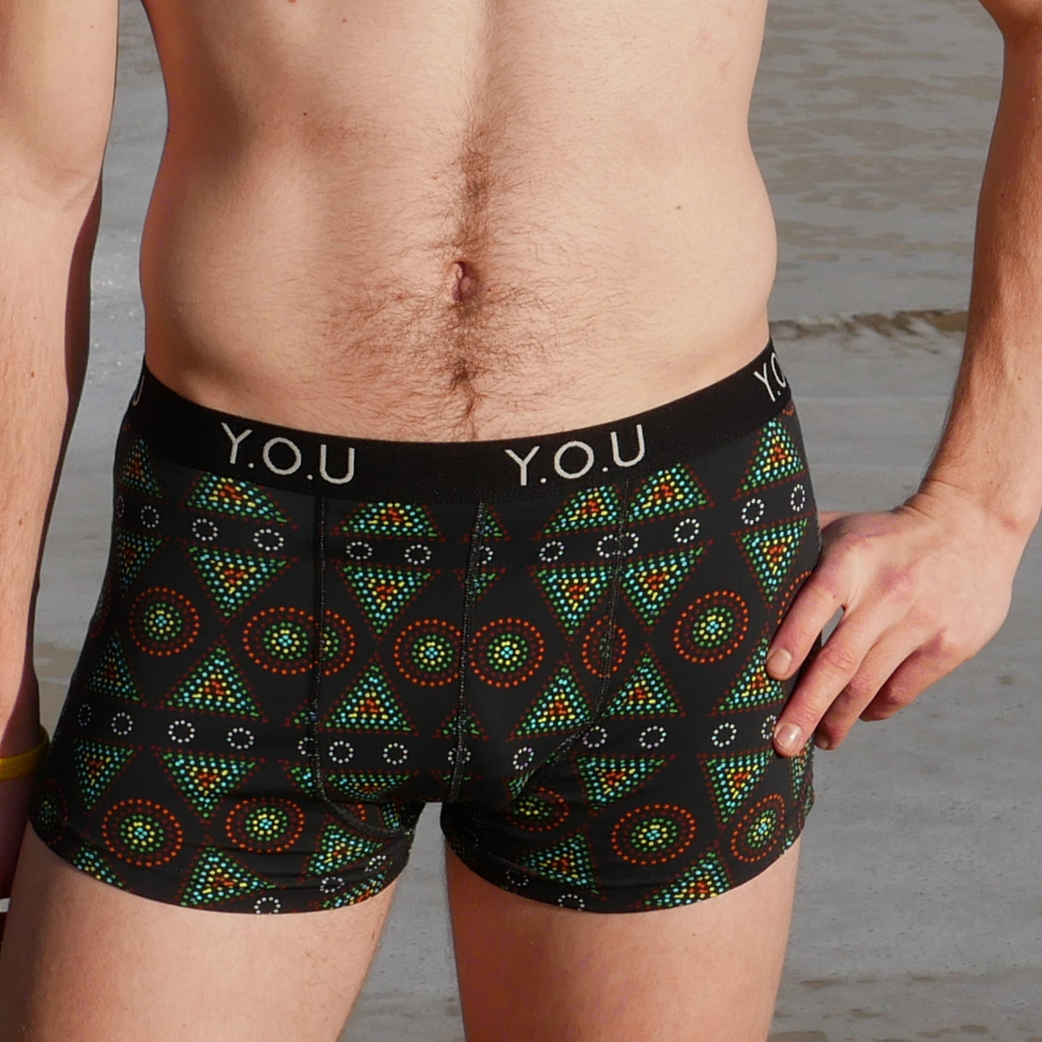 A person stands outdoors wearing black men's hipster trunks with geometric red, green, and orange patterns. The waistband reads "Y.O.U." in white letters. The person's left hand rests on their hip, and part of an arm is visible on their right side.