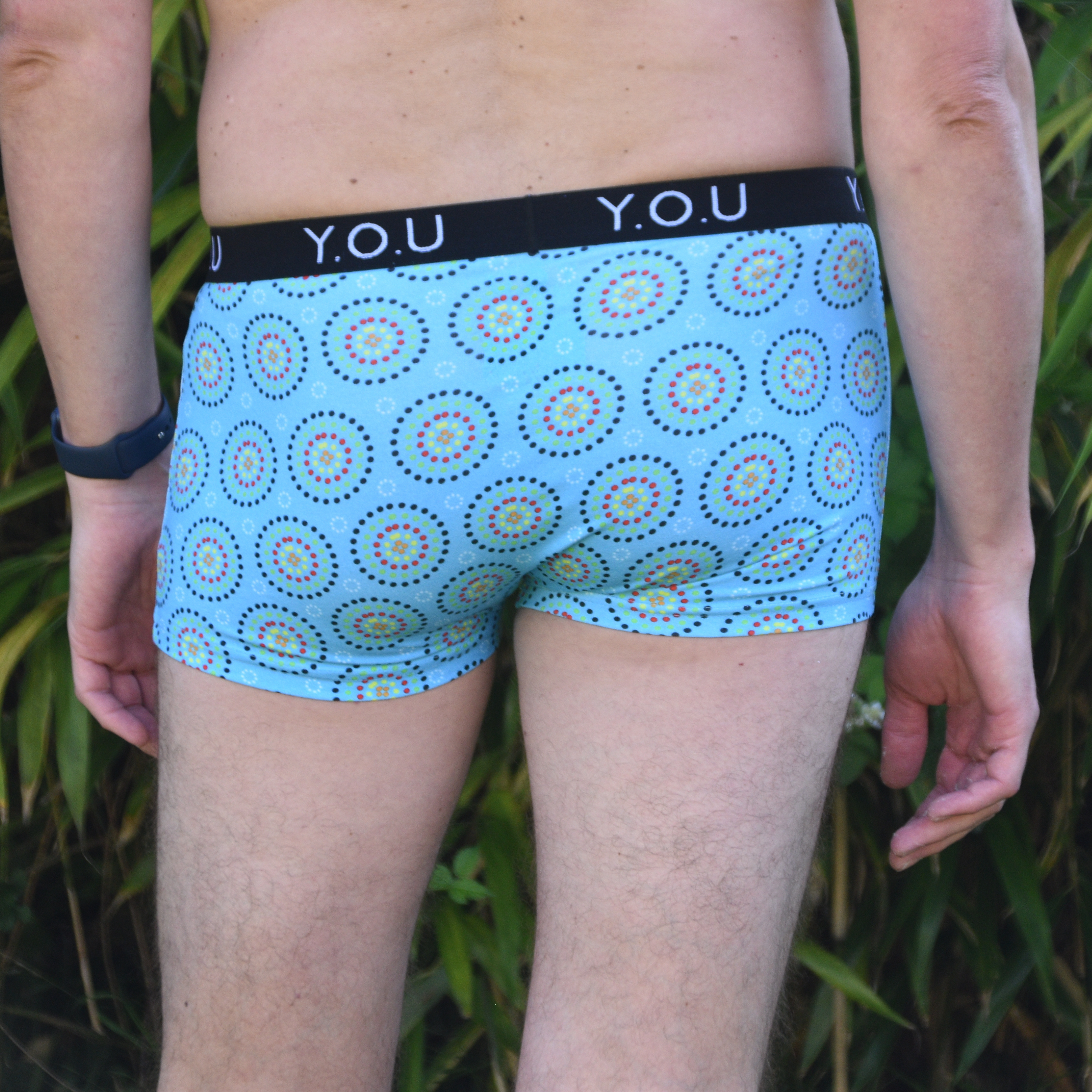 A person stands outdoors, facing away from the camera, wearing light blue men’s hipster trunks with a colourful circular pattern. The waistband reads "Y.O.U." in white letters. The person is near green plants and is wearing a dark wristband on their left wrist.