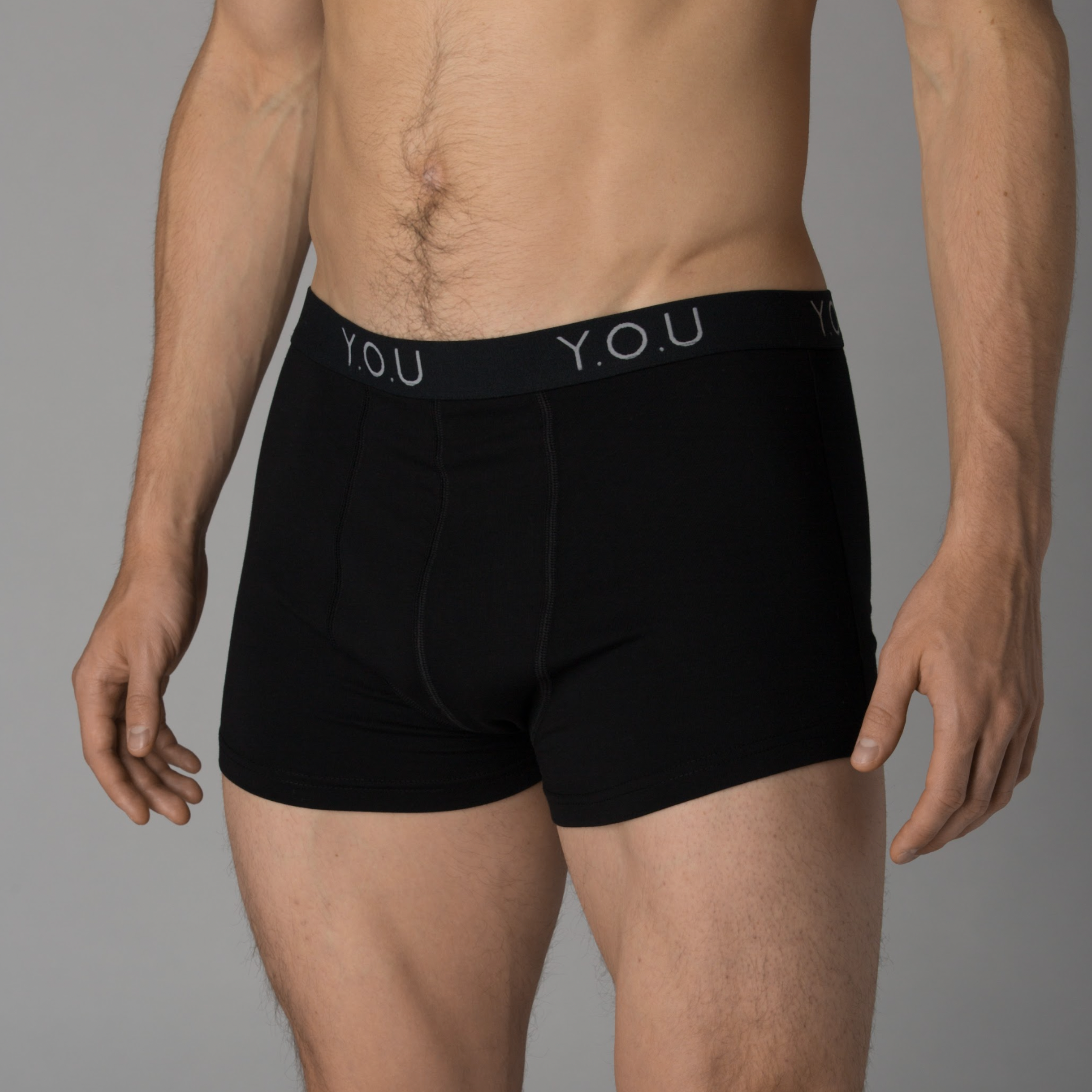 A man is modeling black hipster trunks with "Y.O.U" on the waistband. His upper body is shirtless, and the photo is taken against a plain grey background.