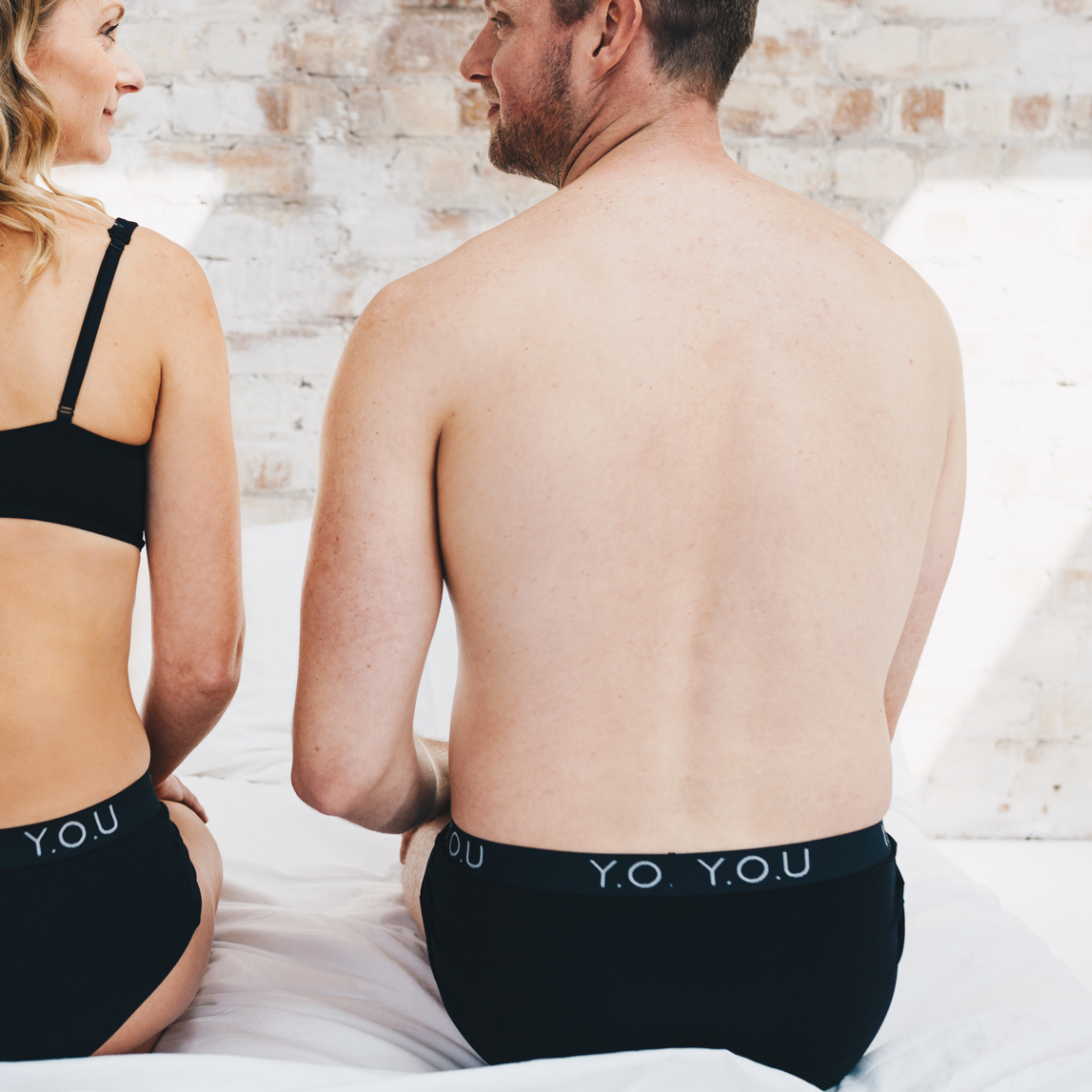 A woman and a man sit on a white bed, facing away from the camera. Both wear black underwear. The man's black hipster trunks have text that reads "Y.O.U" on the waistband. The background features a light brick wall.