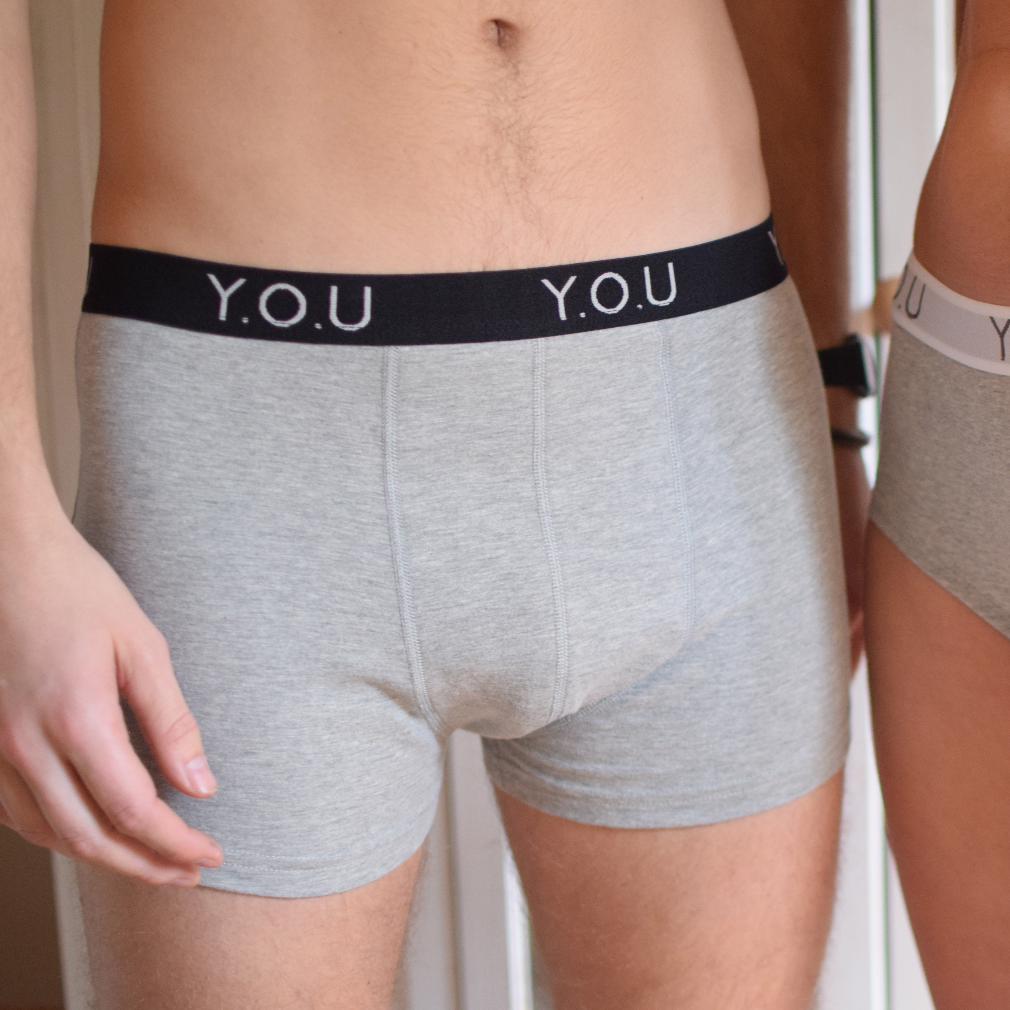 Close-up of two people wearing grey underwear. The person on the left is in grey men's hipster briefs with a black waistband featuring "Y.O.U" in white, made from organic cotton. The person on the right, partially visible, is in similar underwear.
