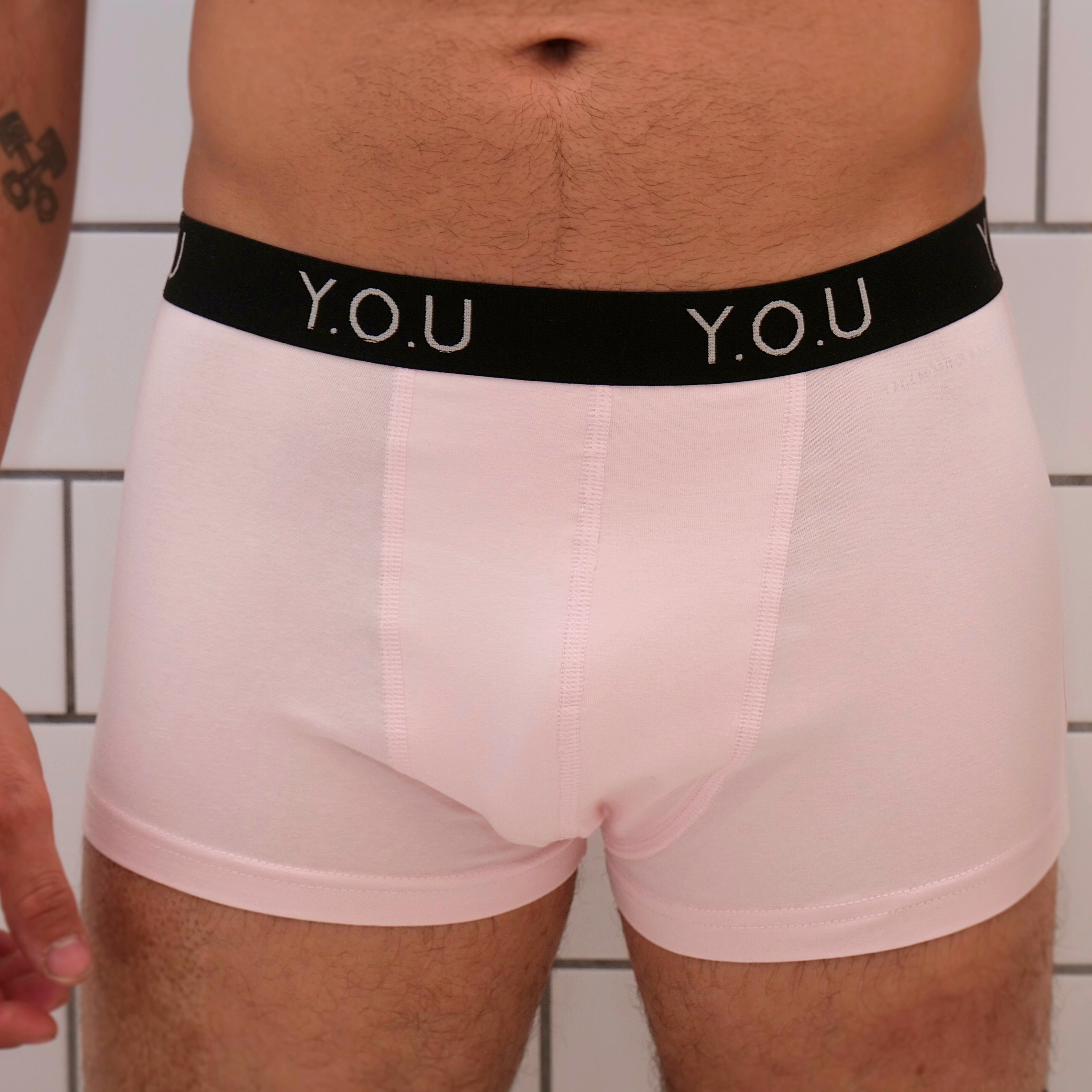Close-up of a person’s lower torso wearing pale pink hipster trunks with a black waistband featuring the text “Y.O.U” in white. The background is a tiled wall. A partial tattoo is visible on the person's right arm.