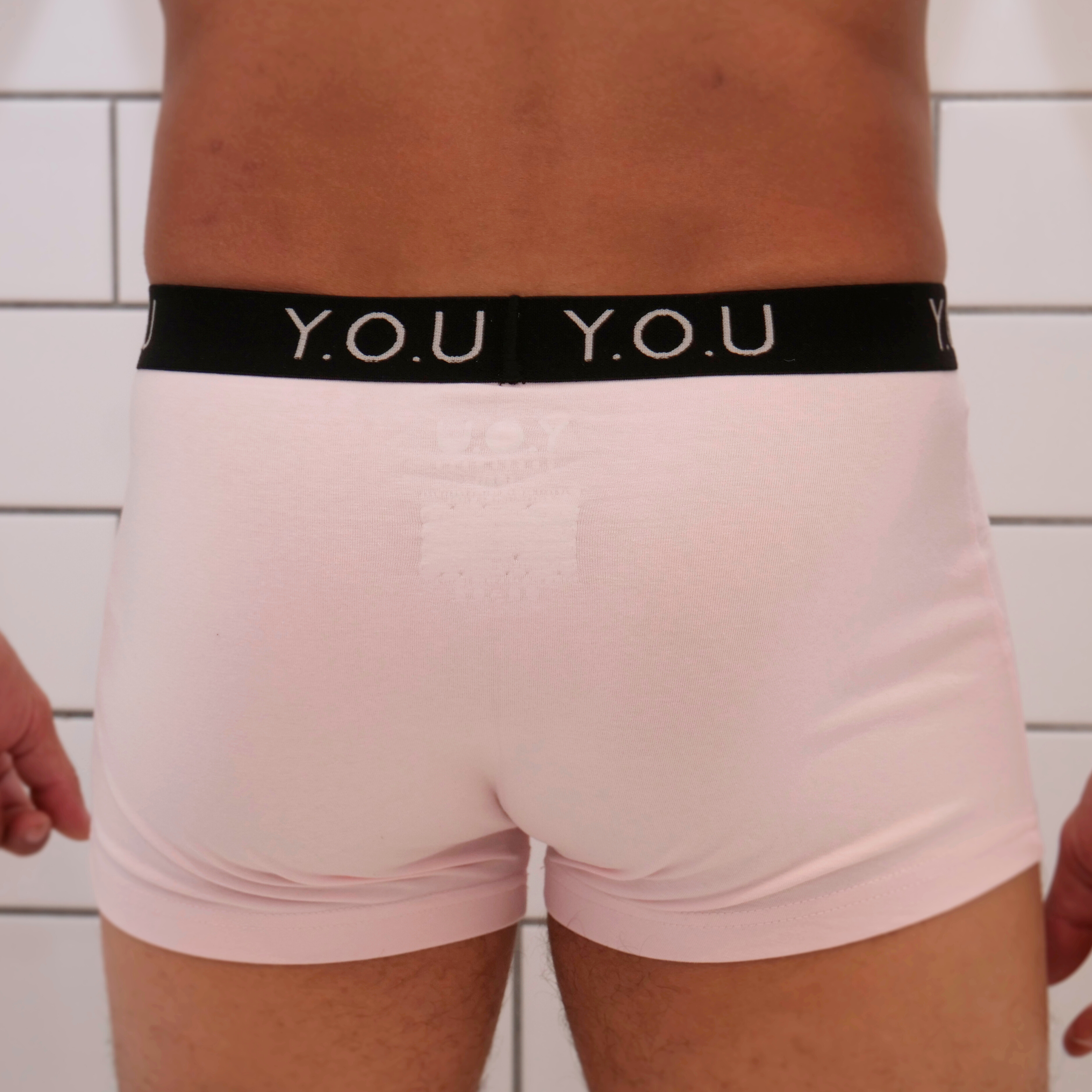 A person wearing light pink hipster trunks with a black waistband that has "Y.O.U" written in white letters across it. The person is standing against a background of white tiled walls, with only their lower body visible in the image.