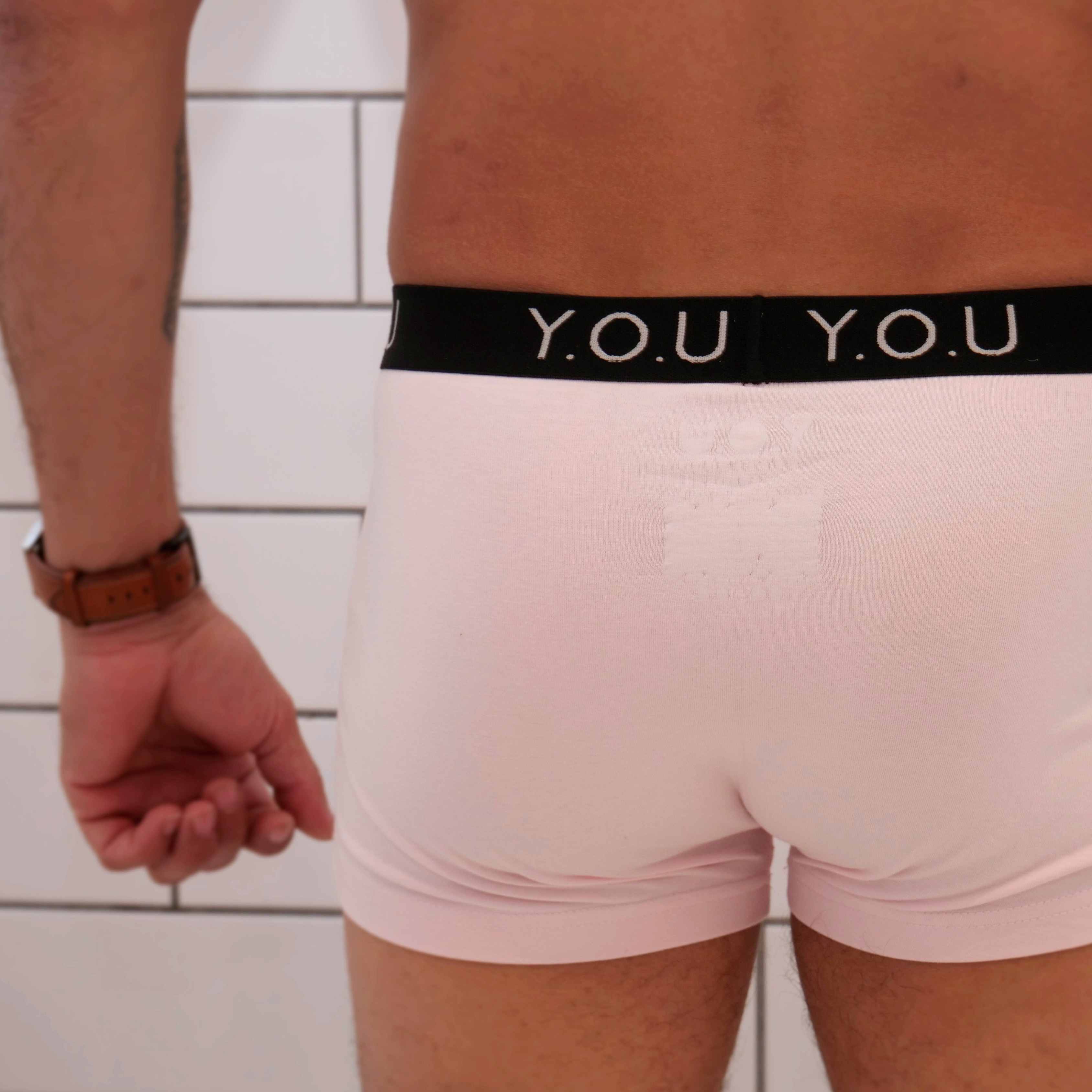 A close-up of a person from behind wearing light pink men's hipster trunks with a black waistband bearing the letters "Y.O.U." 