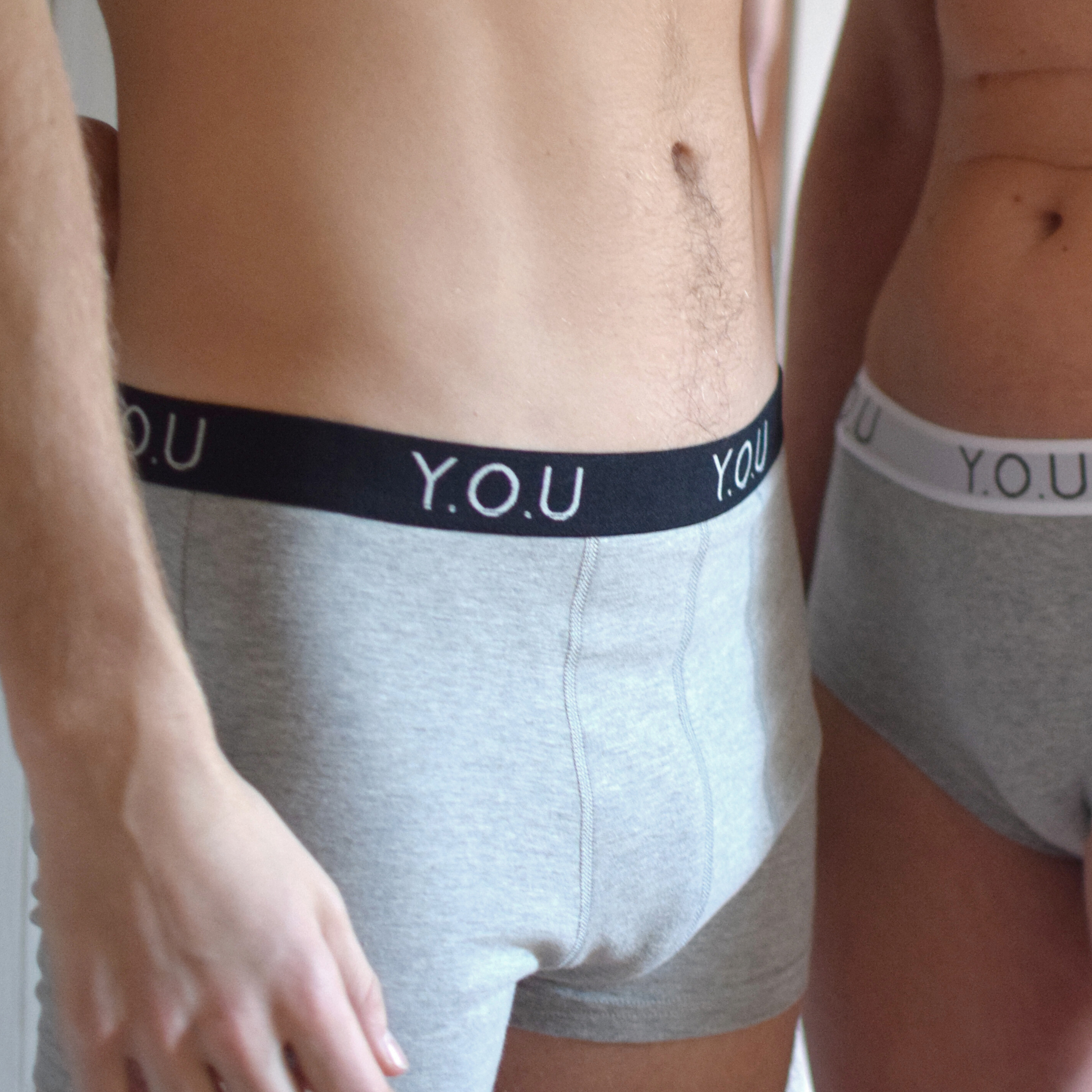 Close-up of two individuals wearing light grey underwear with black waistbands showing the text "Y.O.U" in white. The focus is on the torso and waist area from a side angle, with partial views of their arms and upper thighs, highlighting these  men's hipster trunks.