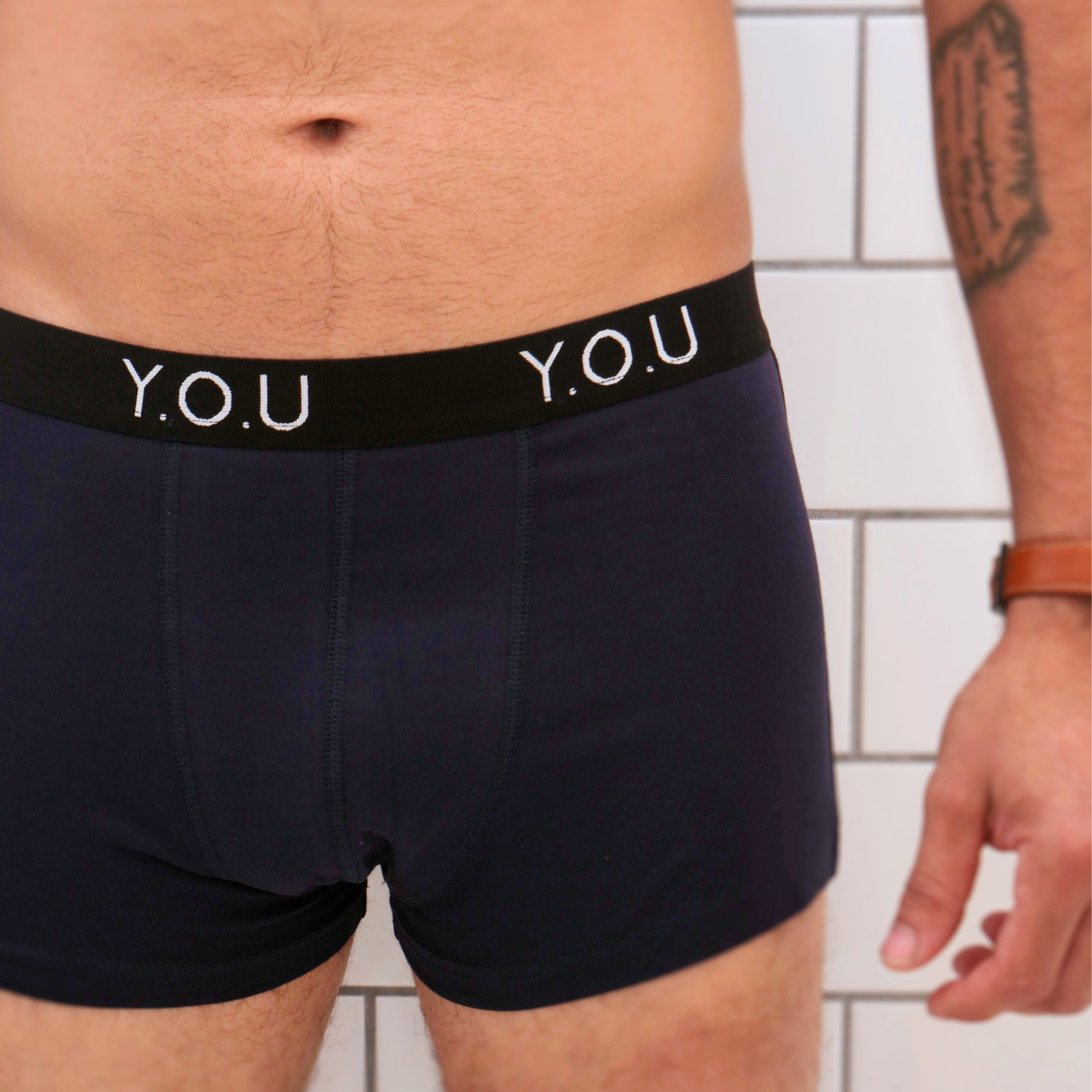 Close-up of a person wearing navy blue men’s hipster trunks with a black waistband that has the letters "Y.O.U" printed on it. The person has a tattoo on their left arm and is wearing a brown wristwatch. They are standing against a tiled wall.