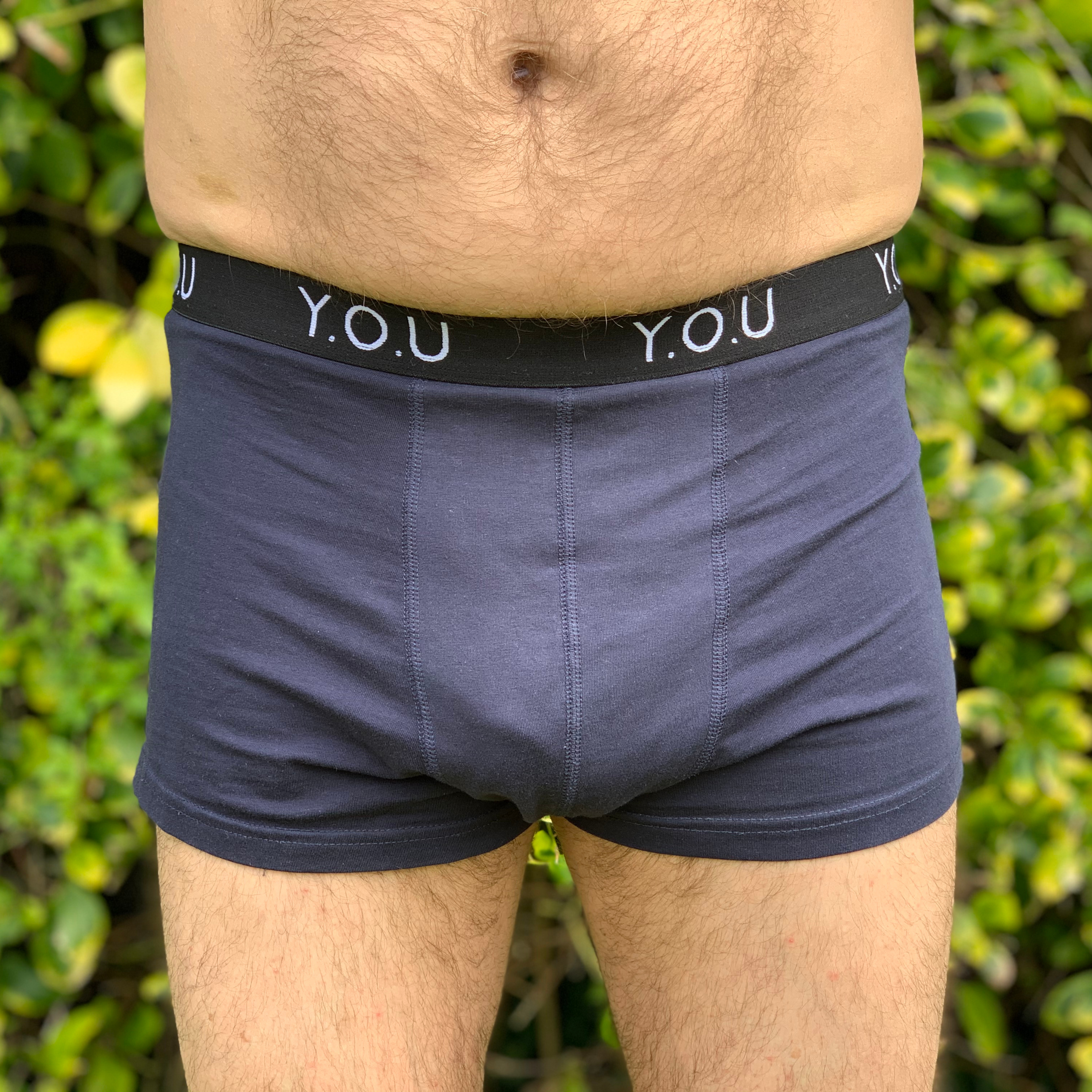 Close-up of a person wearing navy blue men’s hipster trunks with a black waistband that has "Y.O.U" written on it. The person is standing outdoors with green foliage in the background.
