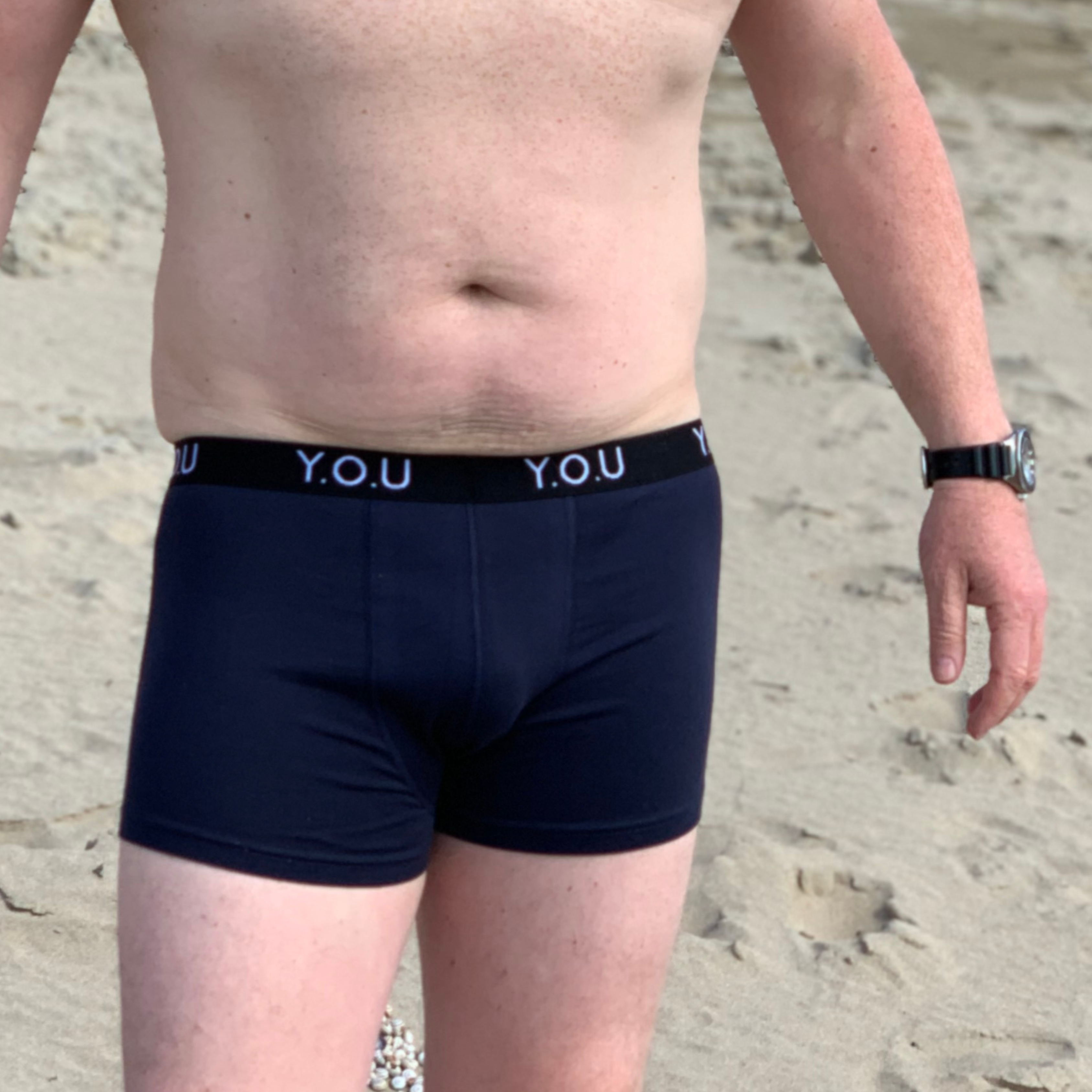 A man with a light complexion wearing navy blue men’s hipster trunks and a watch on his left wrist stands on a sandy beach. The waistband of the trunks has "Y.O.U" written repeatedly, In the background, sand and pebbles are visible but indistinct.