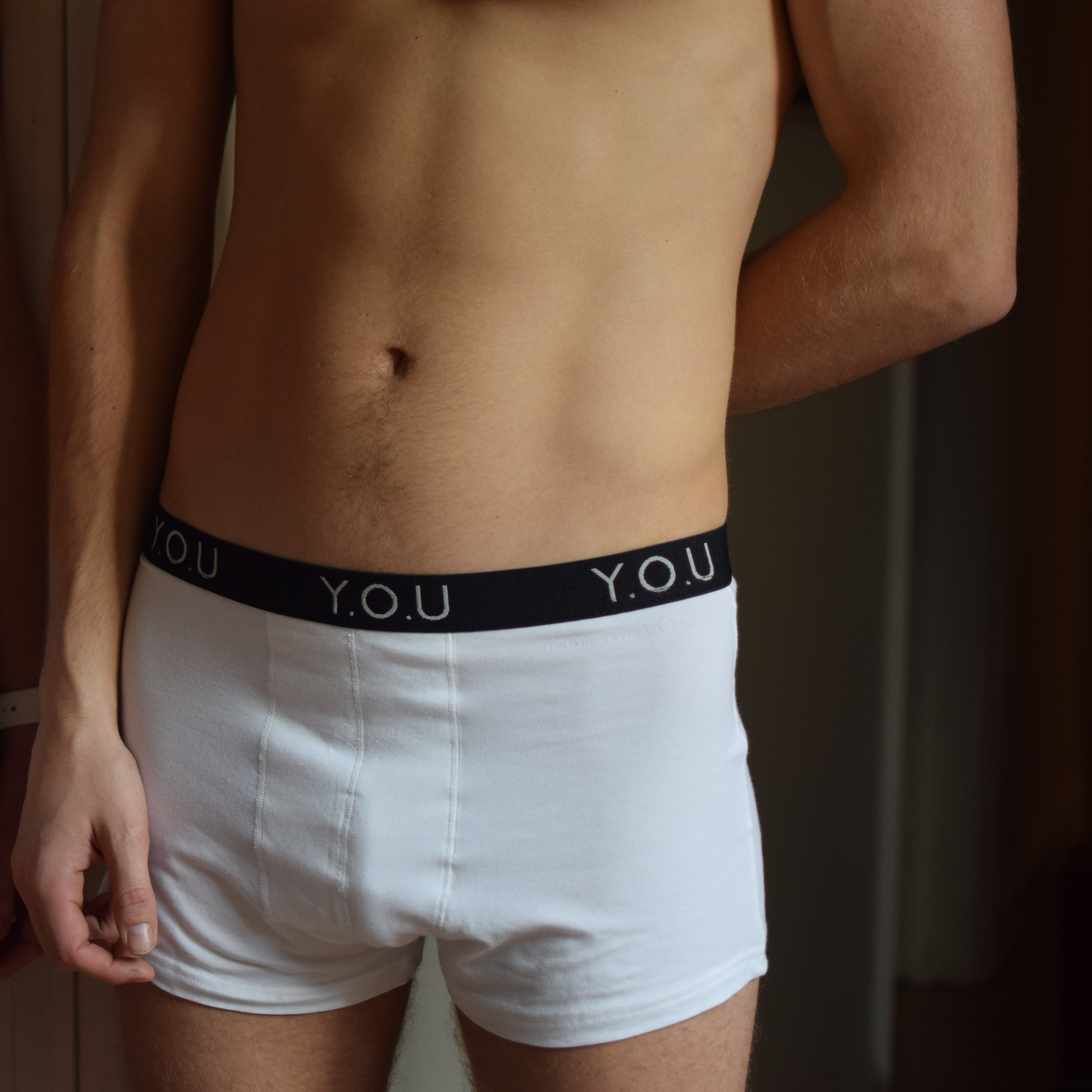 A person is pictured from the neck to the thighs, wearing white men's hipster trunks with a black waistband that has "Y.O.U" printed on it. The individual's hands are hanging naturally by their sides, and the background is slightly blurred.