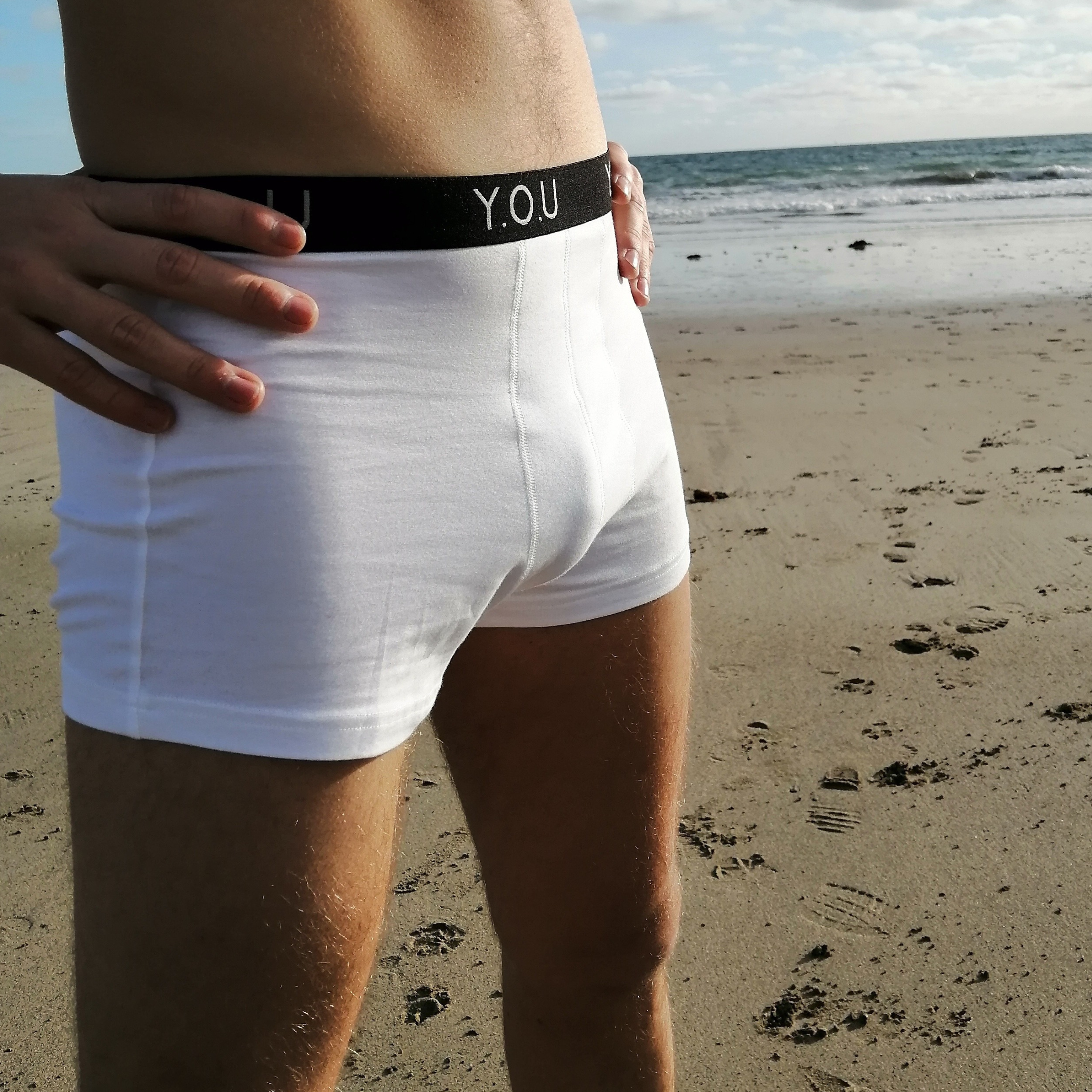 A person standing on a sandy beach near the ocean, wearing a pair of white men’s hipster trunks with a black waistband that has the letters "Y.O.U" on it. The person’s torso and legs are visible, with their hands resting on their hips. The sky is partly cloudy.
