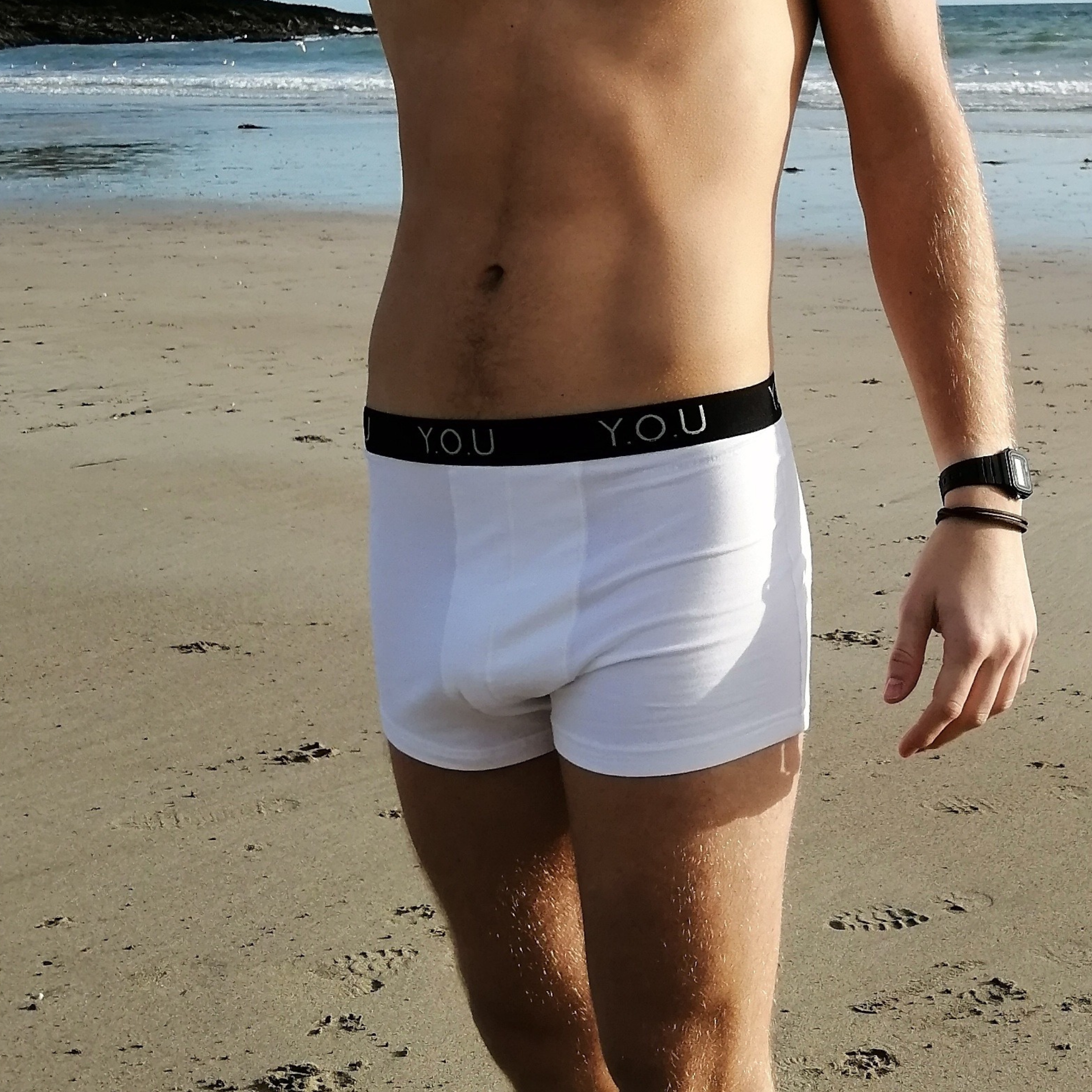 A person wearing white Y.O.U brand men’s hipster trunks and a wristband on their left wrist stands on a sandy beach. Only the lower torso, from mid-chest to mid-thigh, is visible. The beach and waves are in the background.