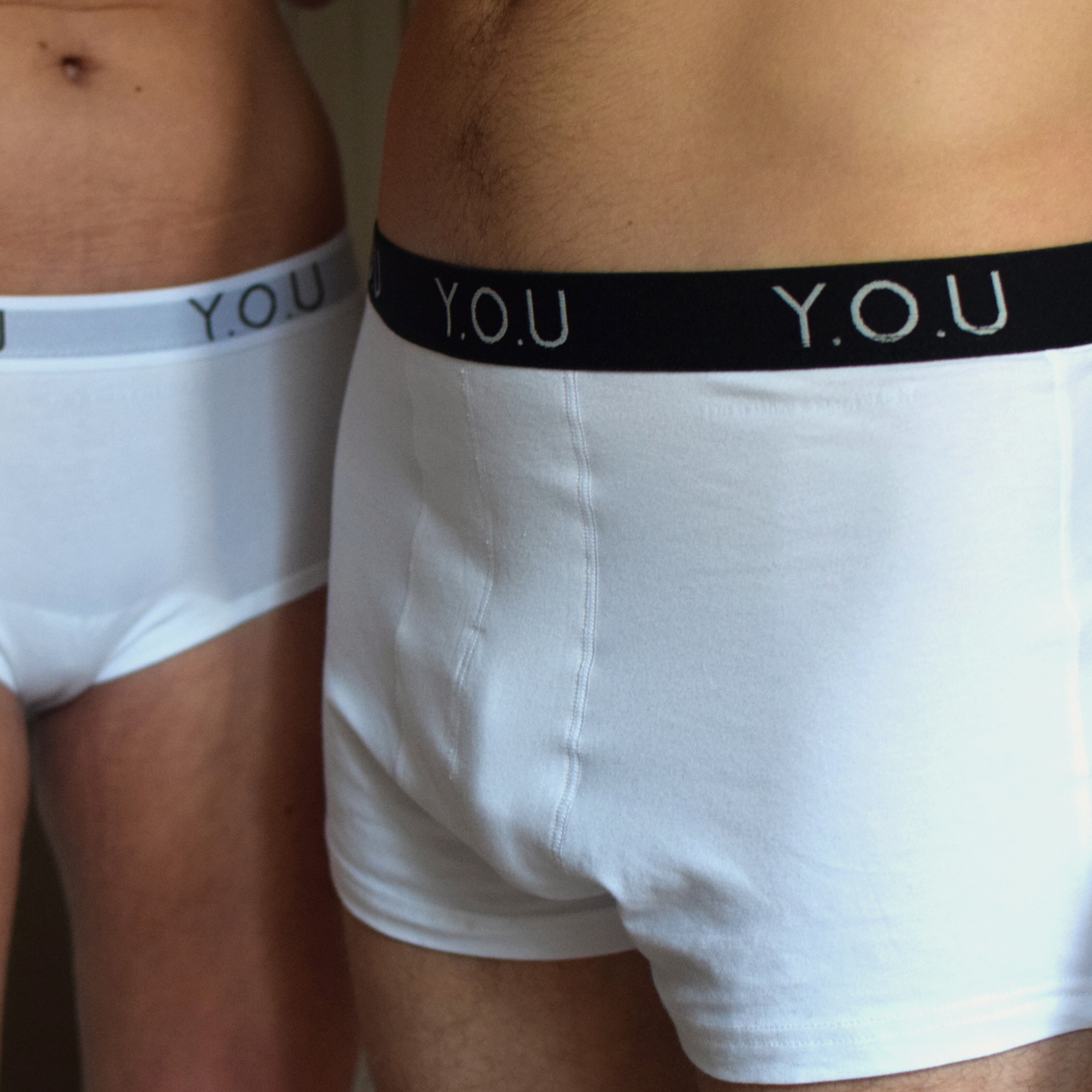 Two individuals are shown from the waist down, wearing white hipster trunks with black waistbands labeled "Y.O.U." The focus is on their midsections as they stand close together.