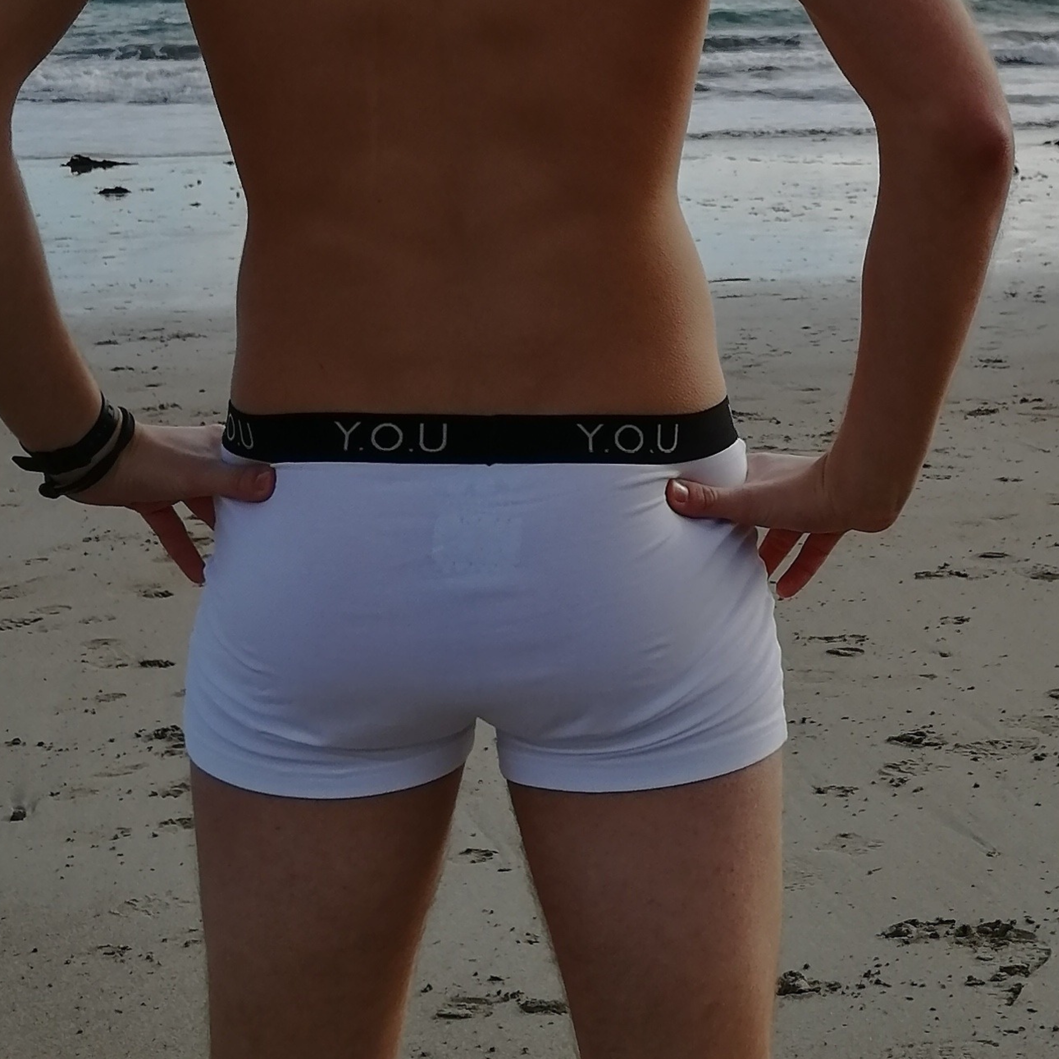 A person with bare upper body stands on a sandy beach facing the ocean, wearing white men’s hipster trunks, with "Y.O.U" written on the waistband. The person has their hands on their hips. Waves are visible in the background.