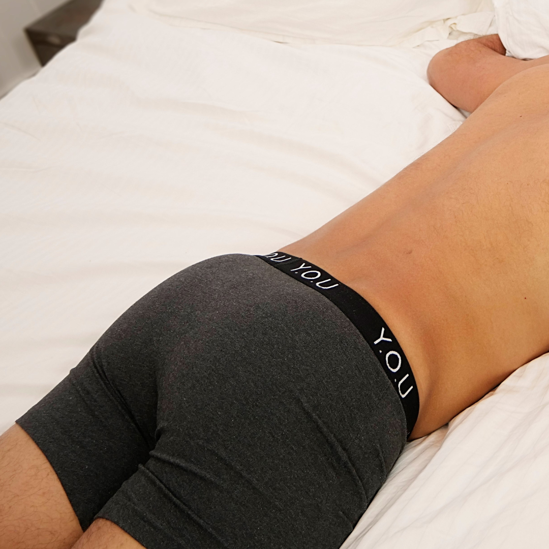A person lies face down on a bed, sporting dark grey men's mid-length trunks that says "YOU" on the waistband. The individual's bare back and part of their arm are visible. The bed is covered with white sheets.