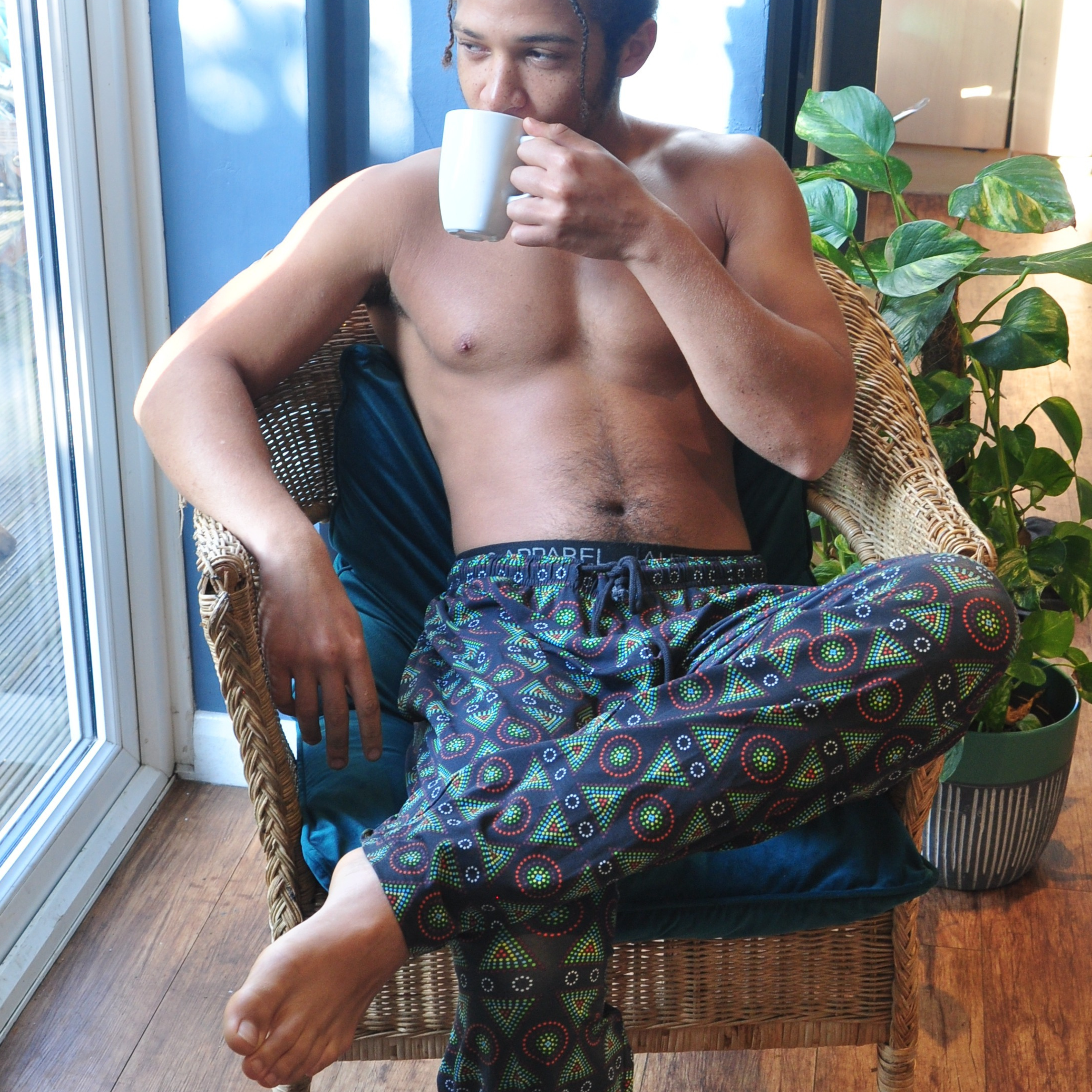 A shirtless person with short hair sits in a wicker chair, drinking from a white mug. They are wearing colorful patterned organic cotton pyjama bottoms and gazing to the side. The scene includes a large window, a leafy green plant, and wooden flooring, giving a cozy ambiance.