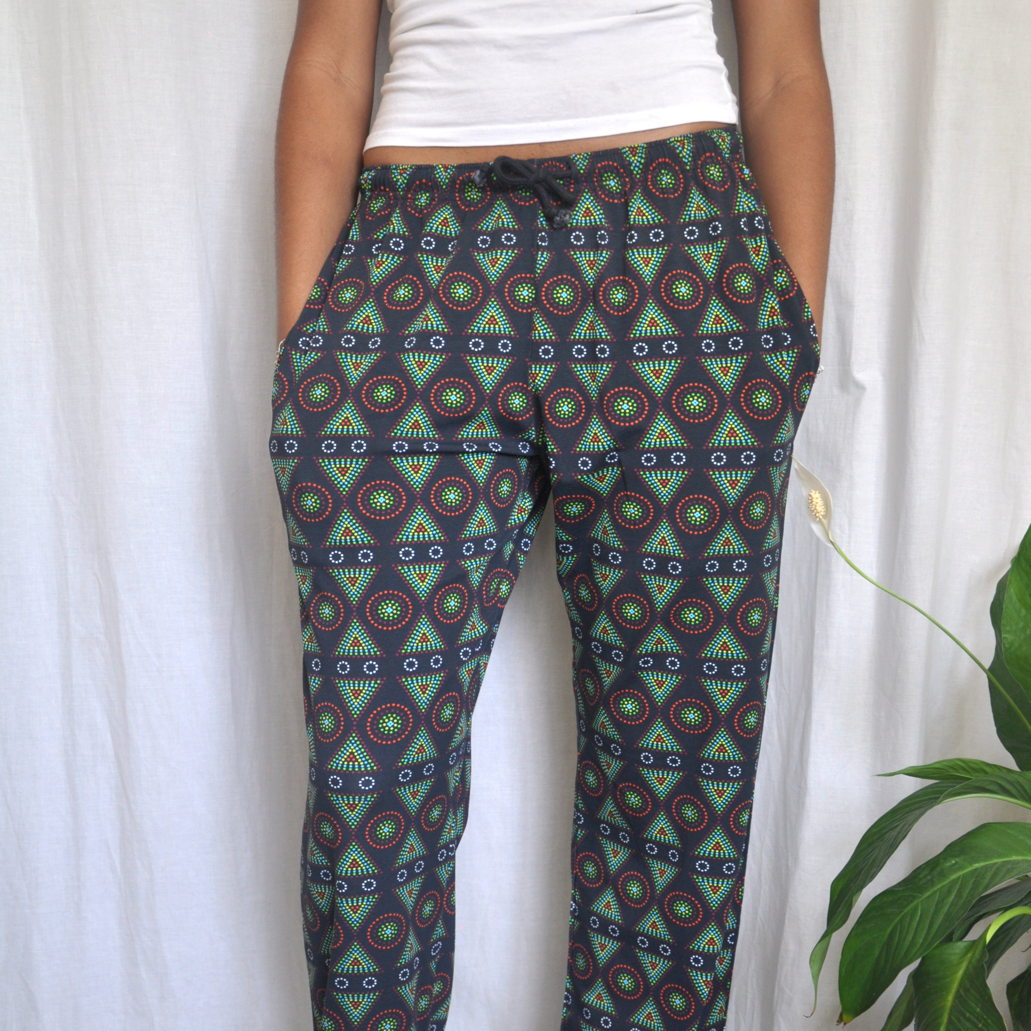 A person is wearing black patterned pyjama bottoms with colorful circular designs and a white shirt. They have their hands in their pockets. The background is a white curtain, and there is a green potted plant on the right side.