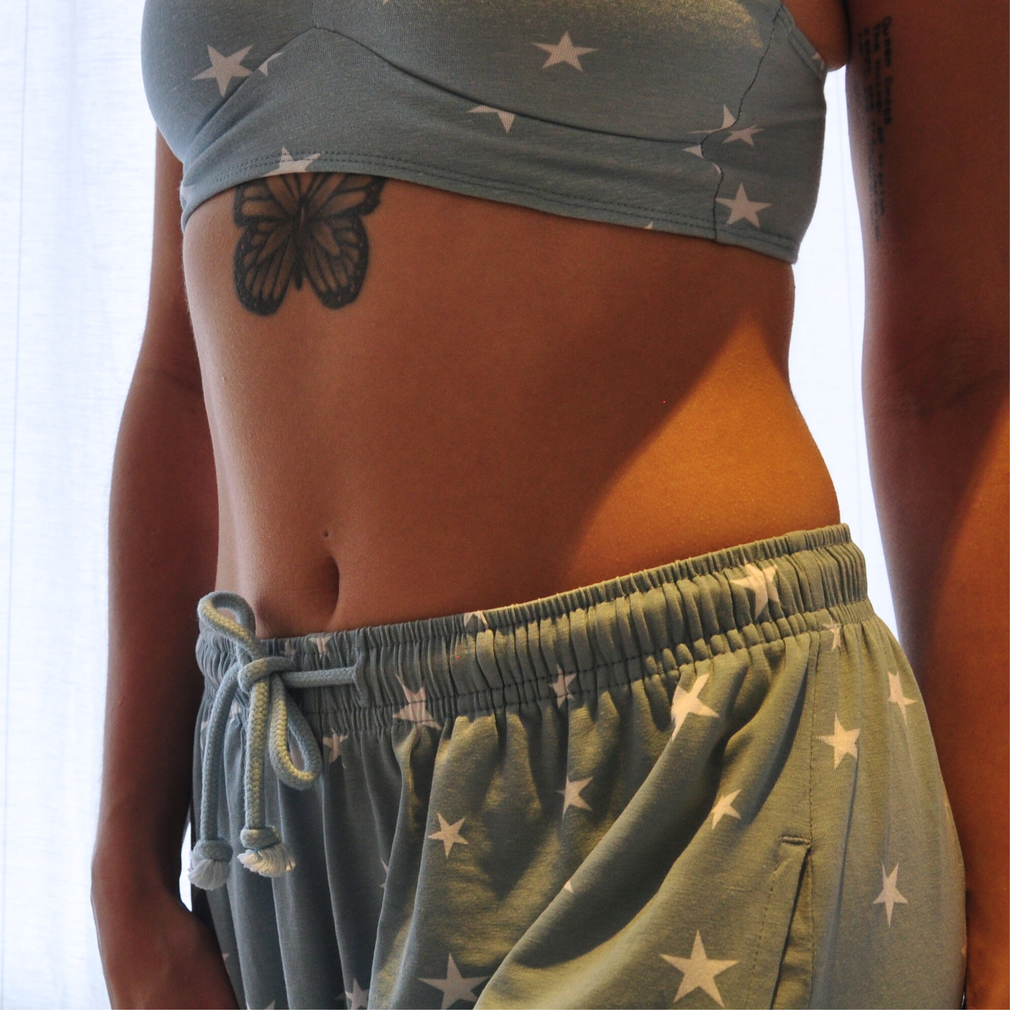 A person stands in front of a light curtain, showing their midsection. They are wearing a light blue bralette with white stars and matching pyjama bottoms. There is a butterfly tattoo under their chest and script text tattooed vertically on their right side.