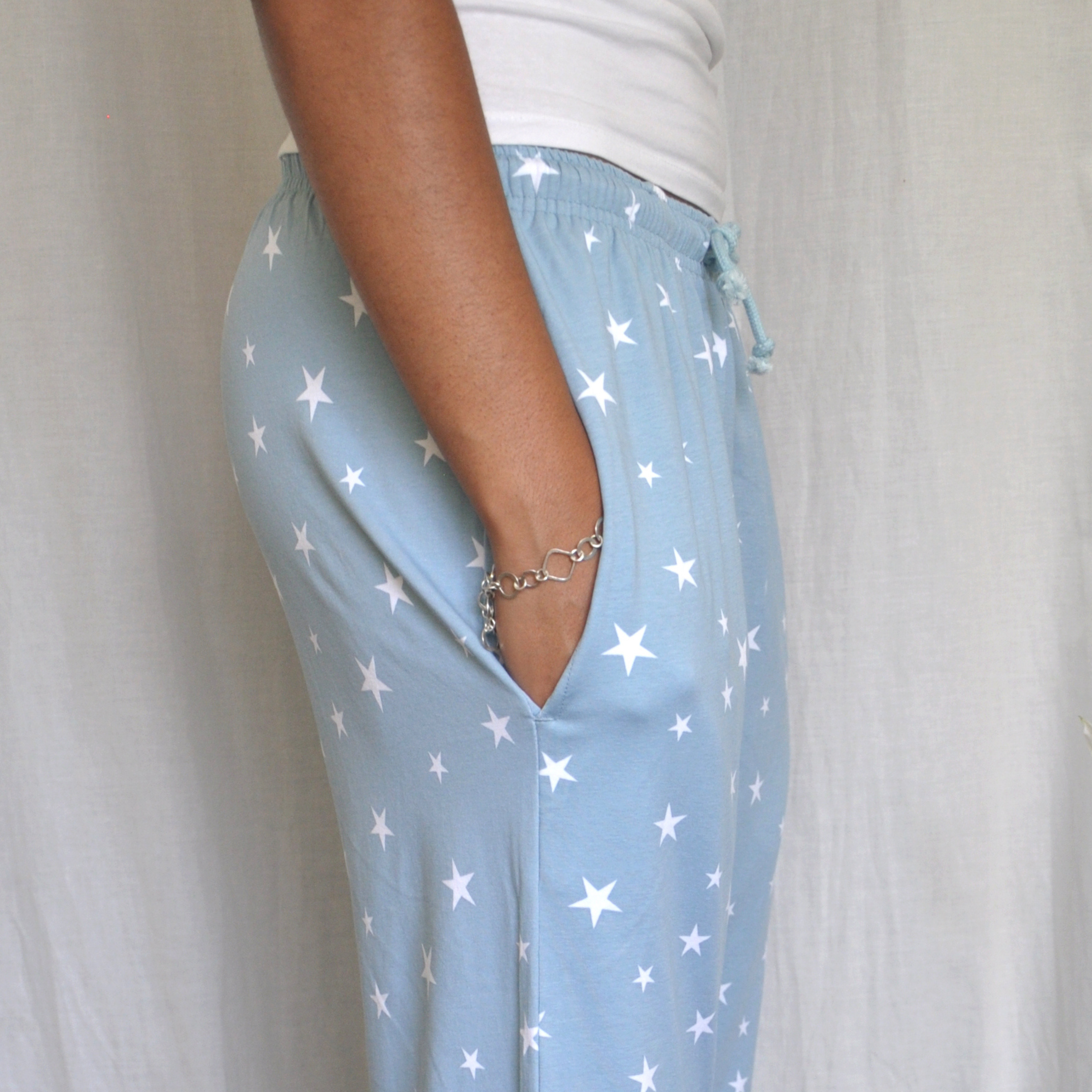 A person is shown from the side wearing light blue pajama pants with white star patterns. They have their hand in the pocket of the pyjama bottoms, which have a drawstring waist. The person is also wearing a white shirt and a silver bracelet. The background is white fabric.