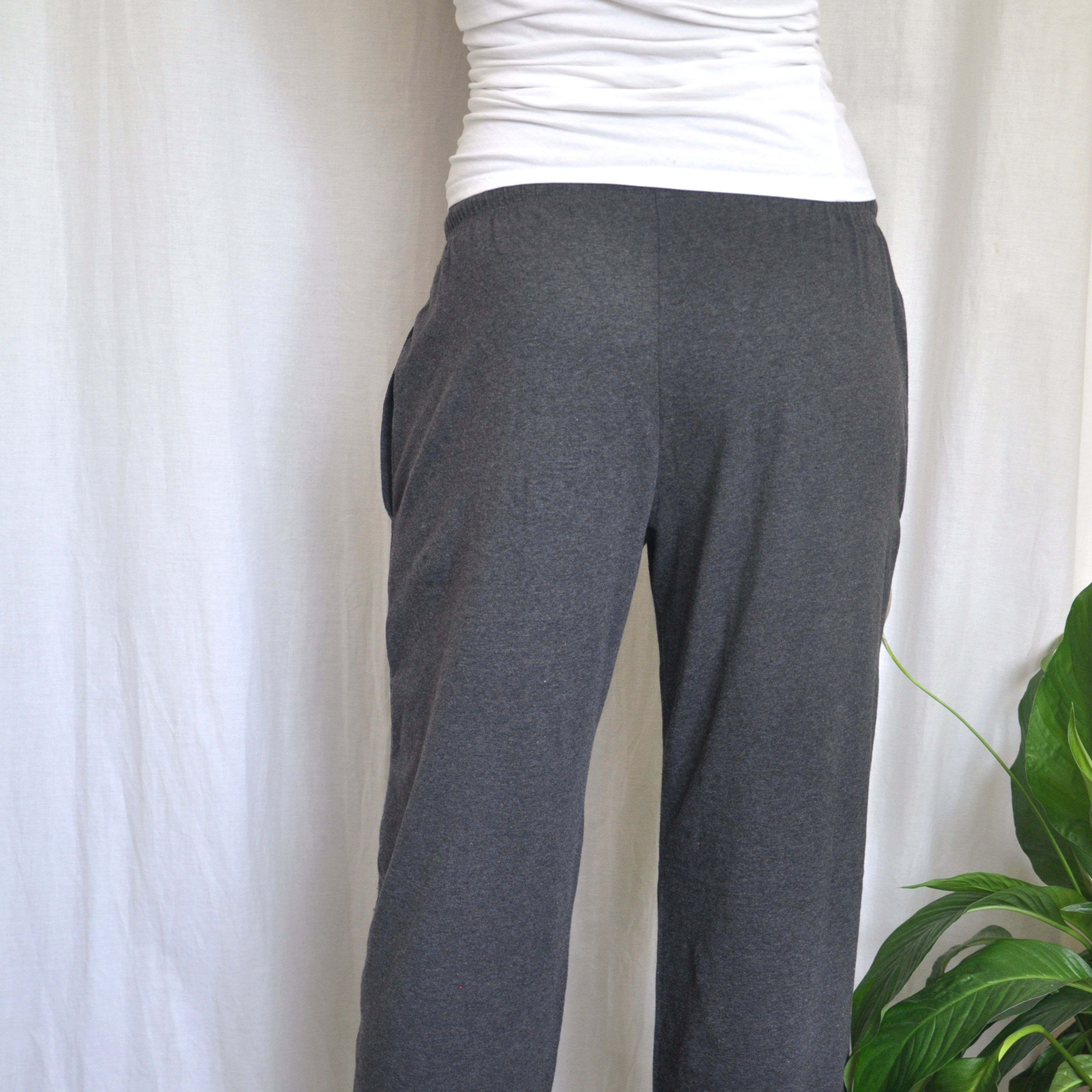 The image shows the lower back view of a person wearing dark grey pyjama bottoms and a white fitted top. The person is standing against a plain white background with a green potted plant visible in the bottom right corner of the frame. 