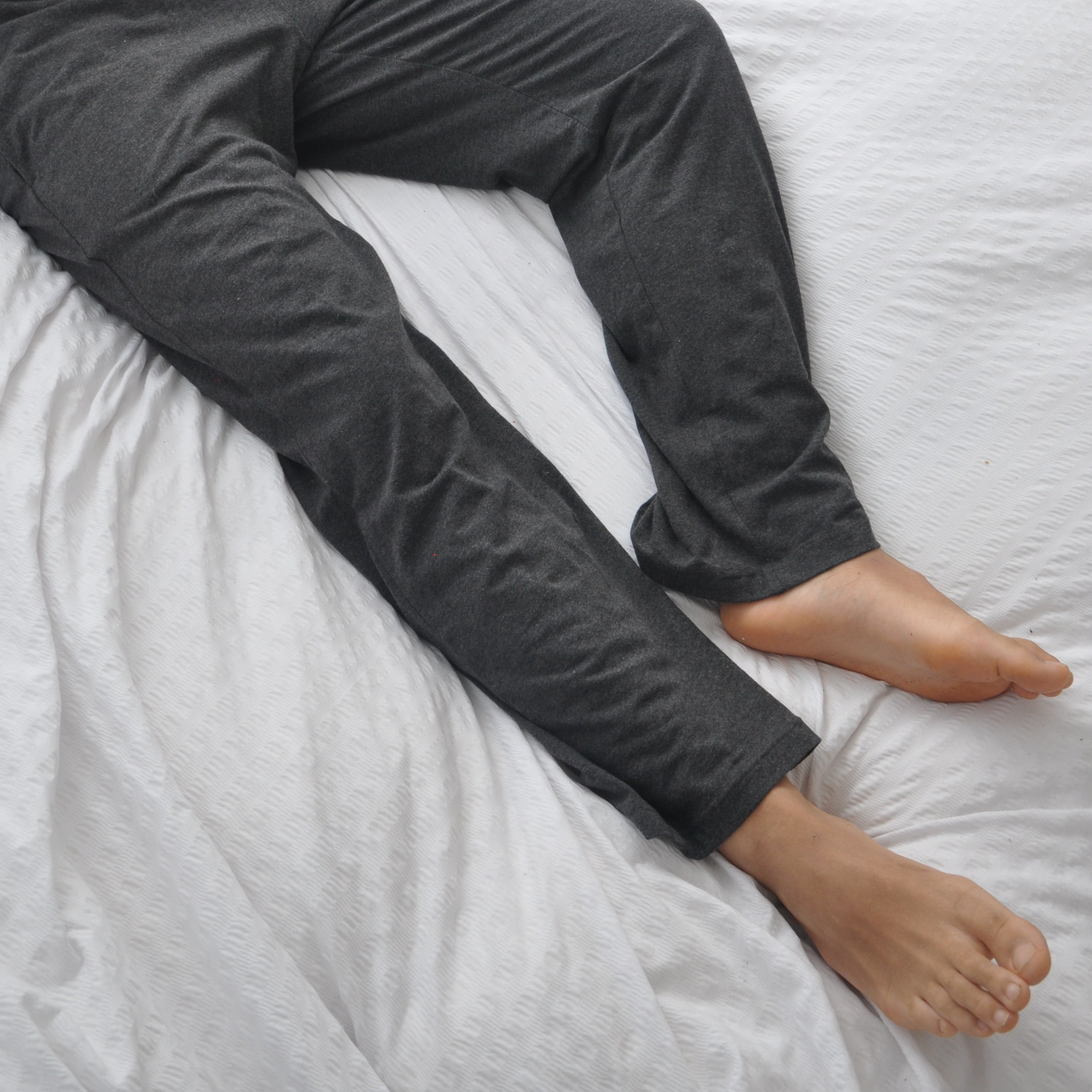 A person wearing dark grey pyjama bottoms is lying on a white bed. Their legs are stretched out, and their bare feet are visible. The bedding has a subtle textured pattern.