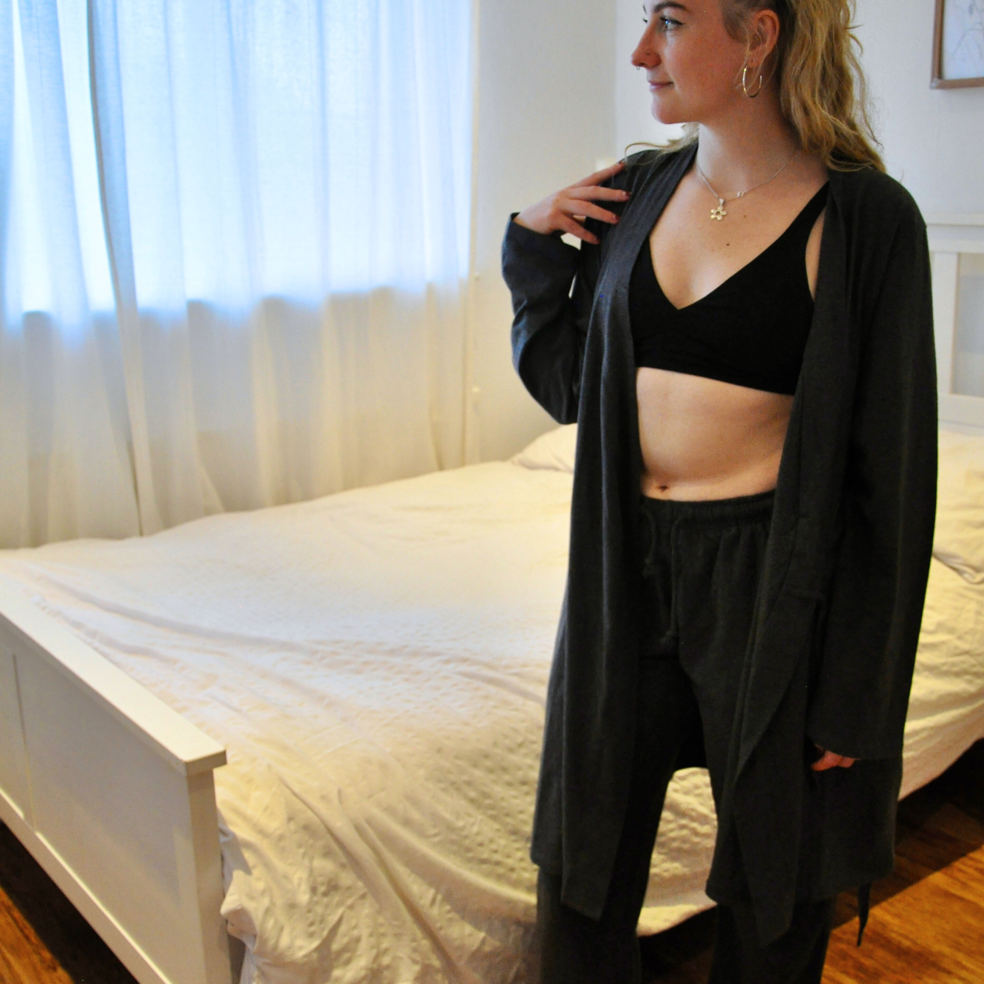 A person stands near a white bed in a room with light streaming through sheer curtains. They are wearing a black bralette, dark grey pyjama bottoms, and an open dark robe, looking to the side with one hand resting near their shoulder. The room features wooden flooring.