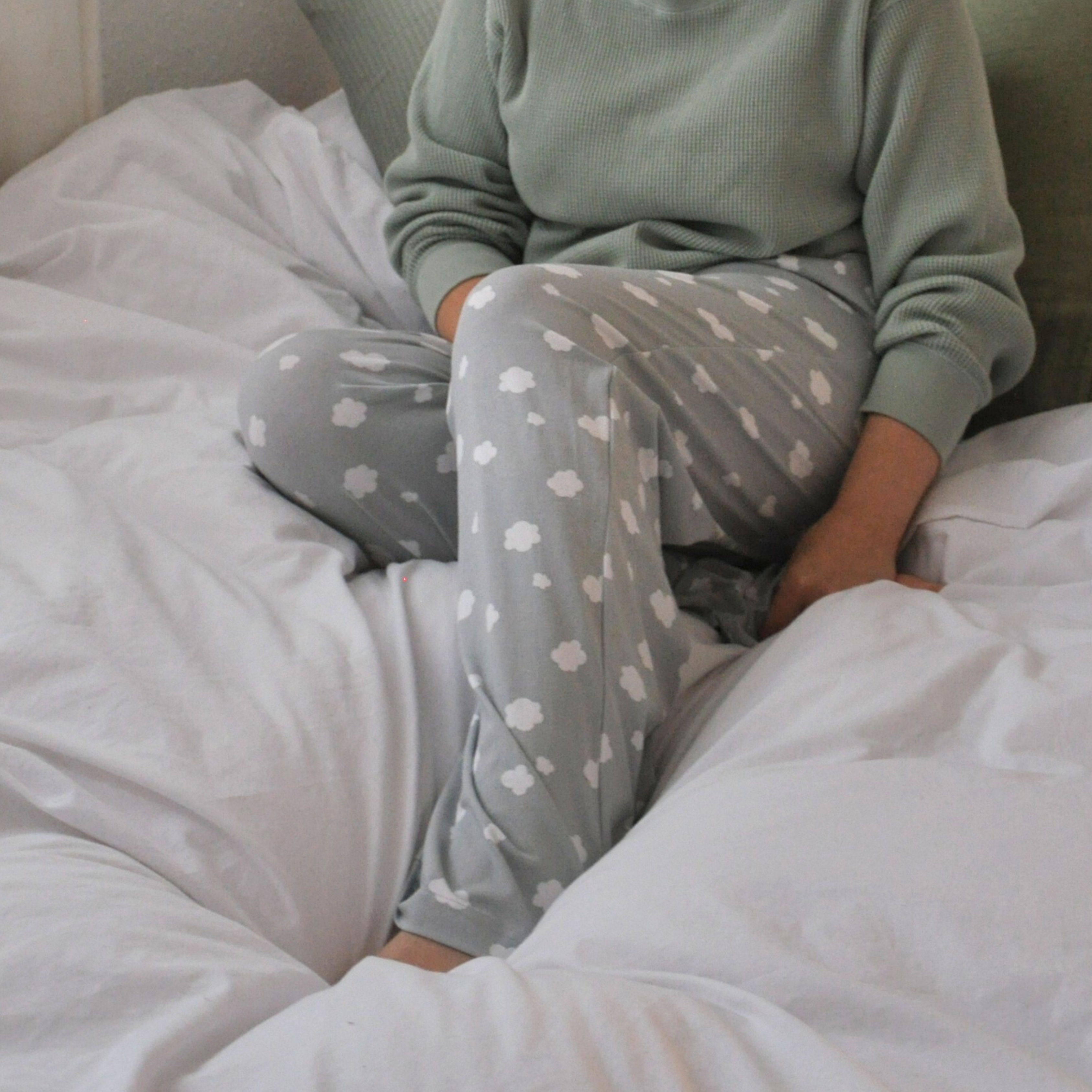 A person with a green sweater and light grey pyjama pants adorned with white cloud patterns is lounging on a bed with white bedding. Only part of the person's torso, arms, and legs are visible in the frame. The background includes pillows and a white wall.