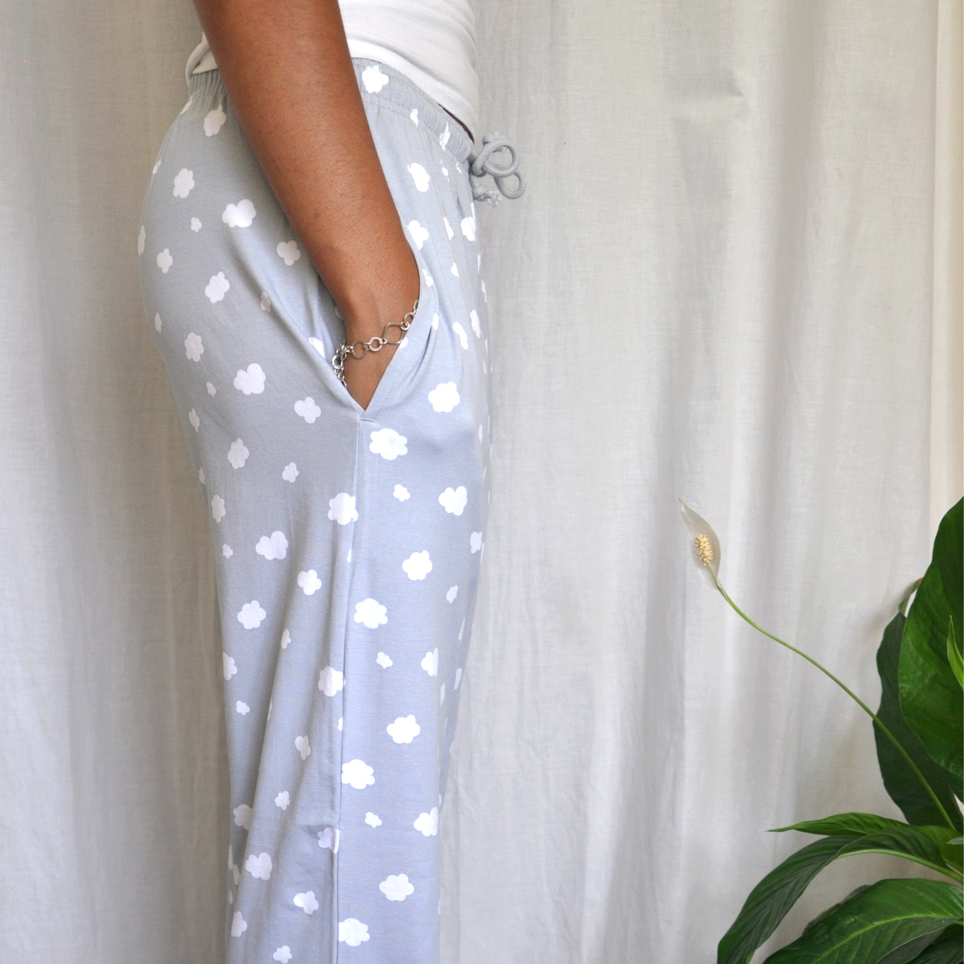 Close-up side view of a person wearing light grey pyjama pants with white cloud patterns. Their left hand is in the pocket, and they are standing in front of a white curtain. A green leafy plant is visible at the bottom right of the image.