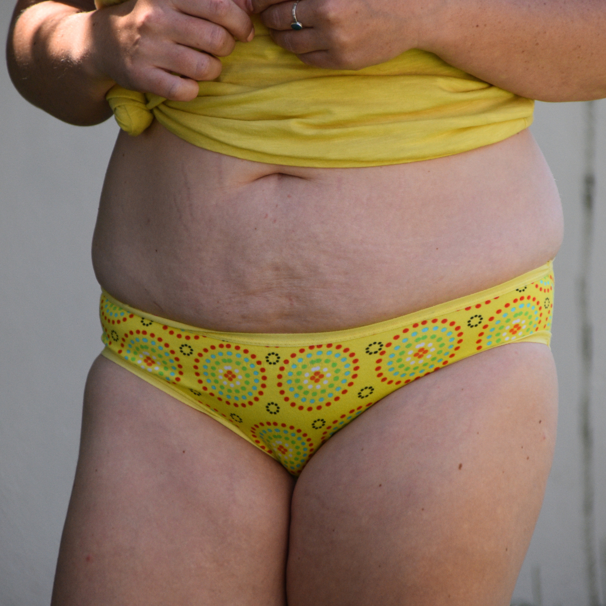 A person is shown from the midsection down, wearing a yellow shirt tied above the waist and yellow underwear with African inspired prints. The person's hands are holding the knot of the shirt.